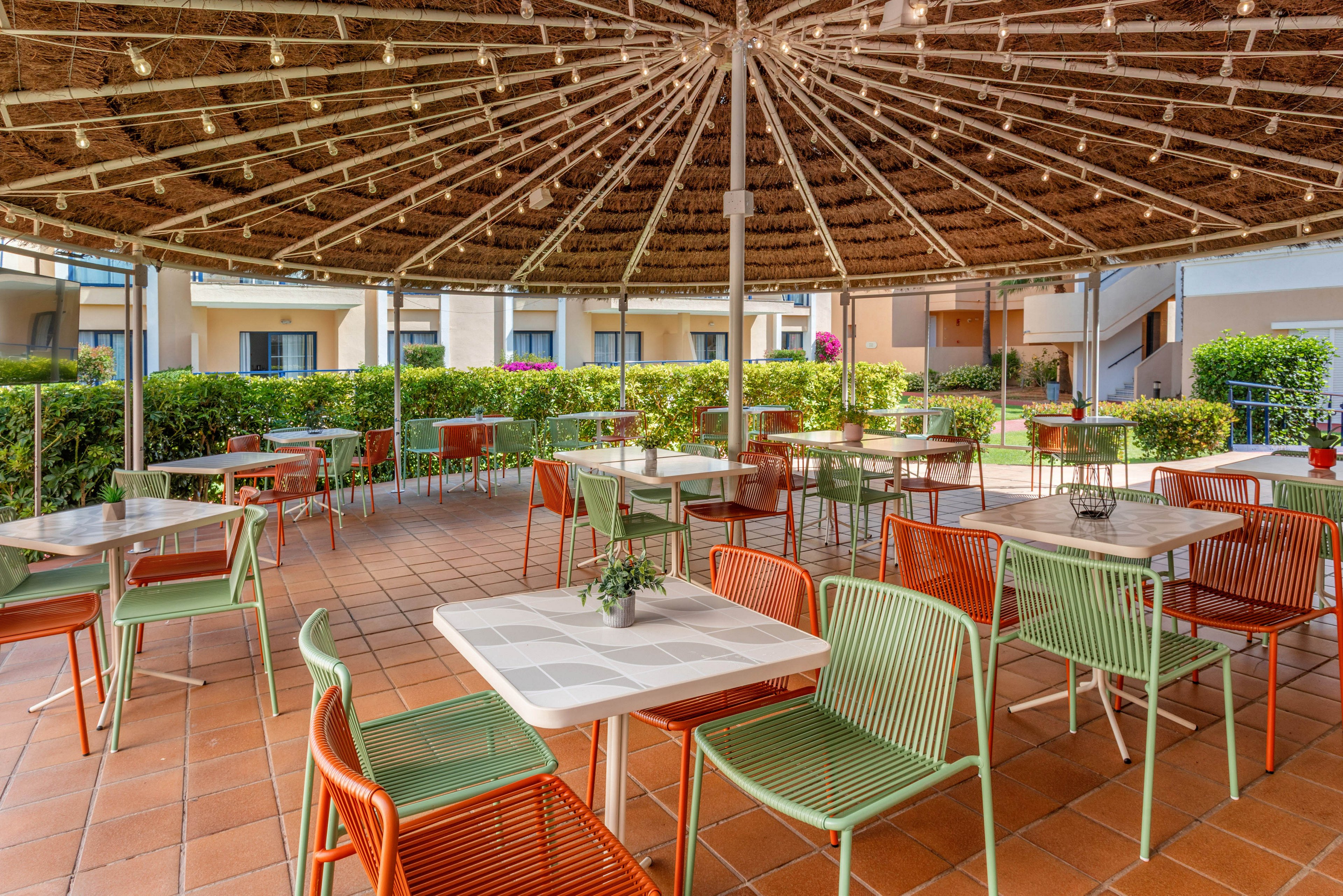 An open-air patio café features neatly arranged tables and colorful metal chairs under a large circular thatched canopy. The tiled floor and surrounding green hedges create a clean, inviting environment. Apartment-style buildings with balconies frame the background, suggesting a hotel or residential complex. Soft natural light and warm tones give the scene a relaxed, summery atmosphere.