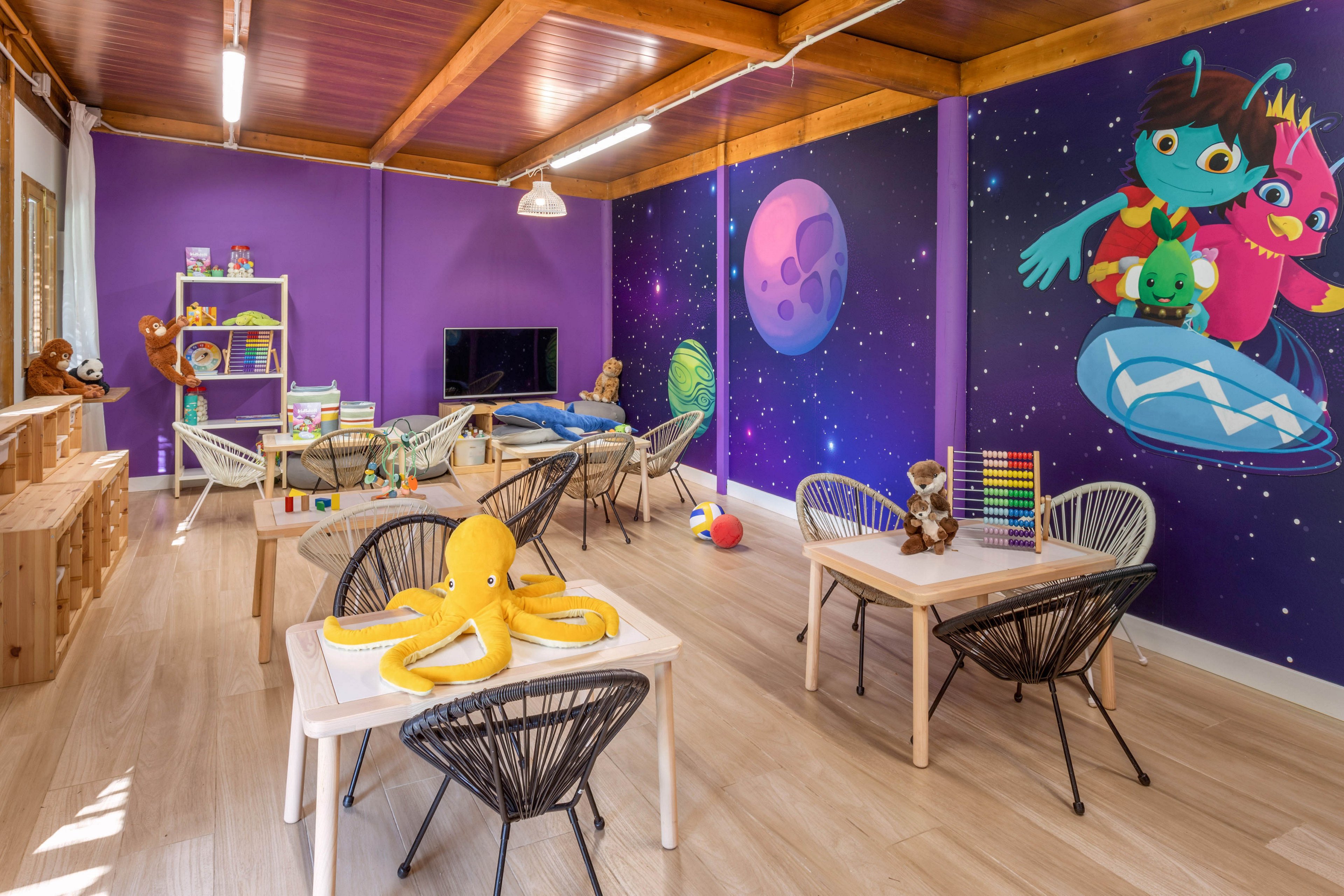 A bright children's playroom features purple walls decorated with a whimsical space-themed mural of cartoon aliens and planets. Small tables and chairs are arranged around the room with toys, books, and stuffed animals neatly displayed. Wooden shelving, a television, and educational toys such as an abacus and stacking blocks add to the playful learning environment. Warm wooden ceilings and light-colored flooring create a cozy, inviting atmosphere for kids.