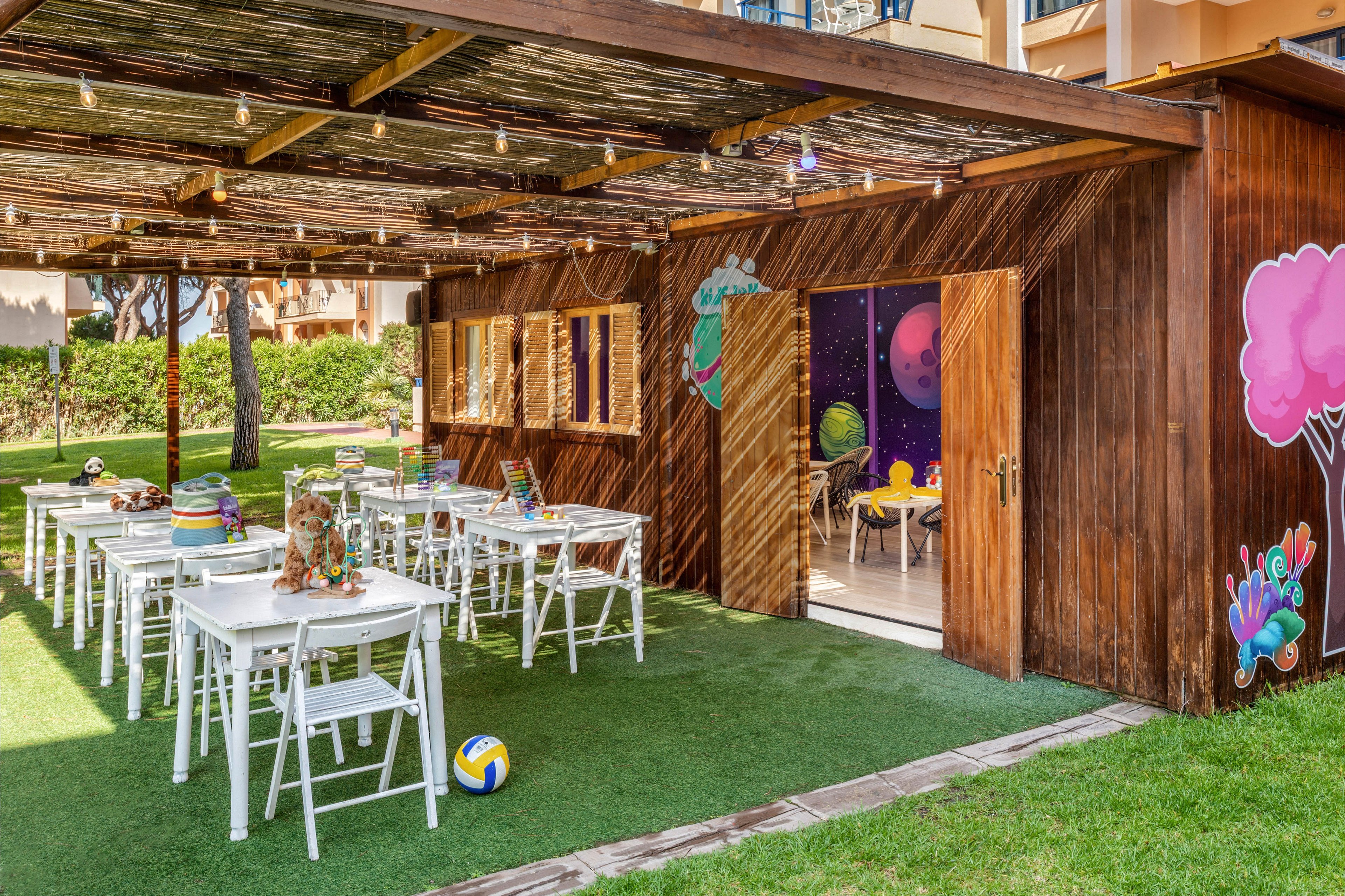 A wooden open-air pavilion is set up with rows of white tables and chairs arranged for a children’s event. The structure opens onto a colorful indoor playroom decorated with space-themed wall art and bright furniture. String lights hang from the rustic roof, and the area is surrounded by green artificial turf and landscaped gardens. A volleyball and various decorations add to the playful, festive atmosphere.