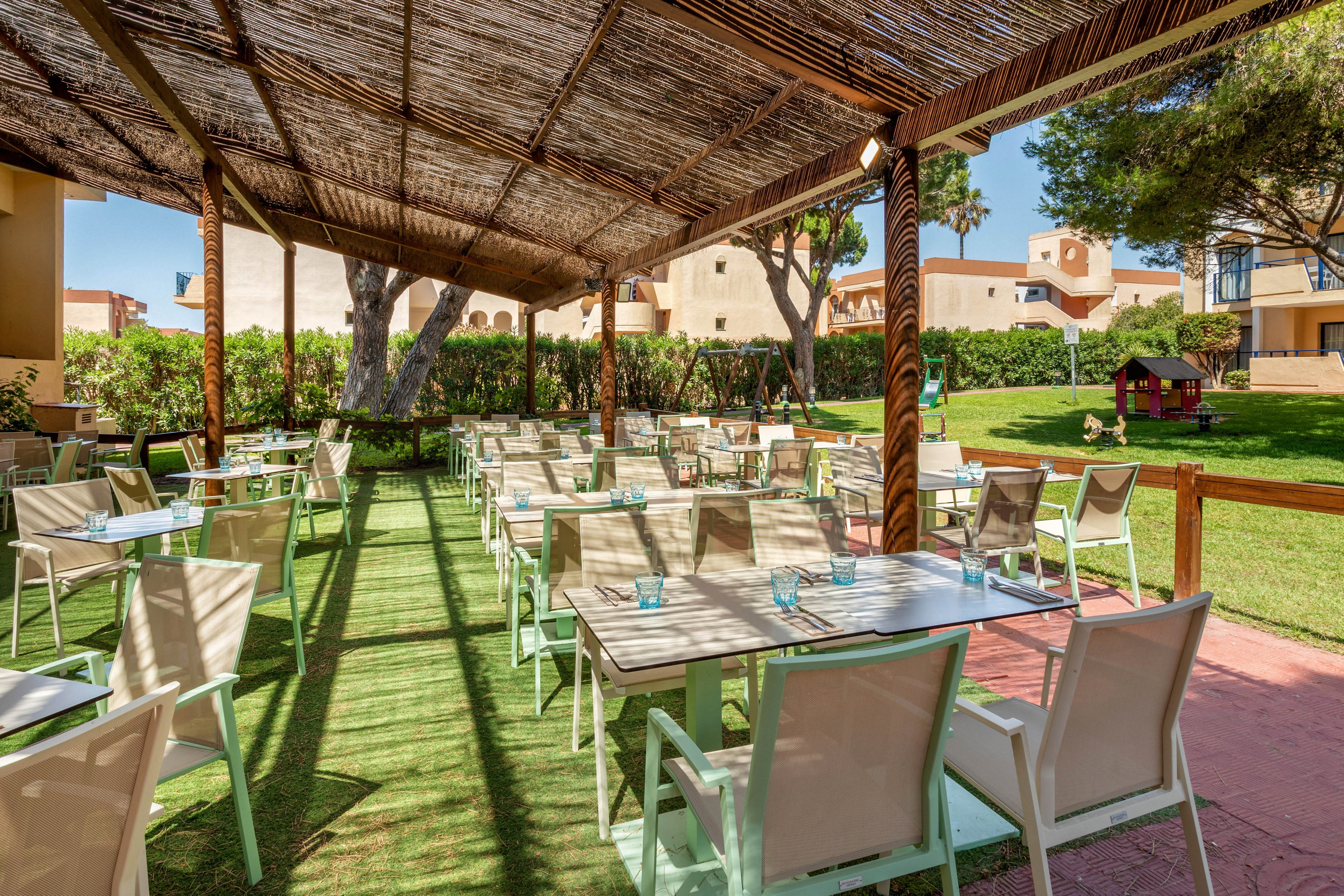 An open-air dining area is set under a rustic pergola with a woven reed roof providing shade. Multiple tables and chairs are neatly arranged on a green artificial grass floor, each table set with small blue glasses. The terrace overlooks a landscaped garden with trimmed hedges, trees, and a small children's playhouse. In the background, warm-toned resort buildings and balconies are visible under a clear sunny sky, creating a relaxed vacation atmosphere.
