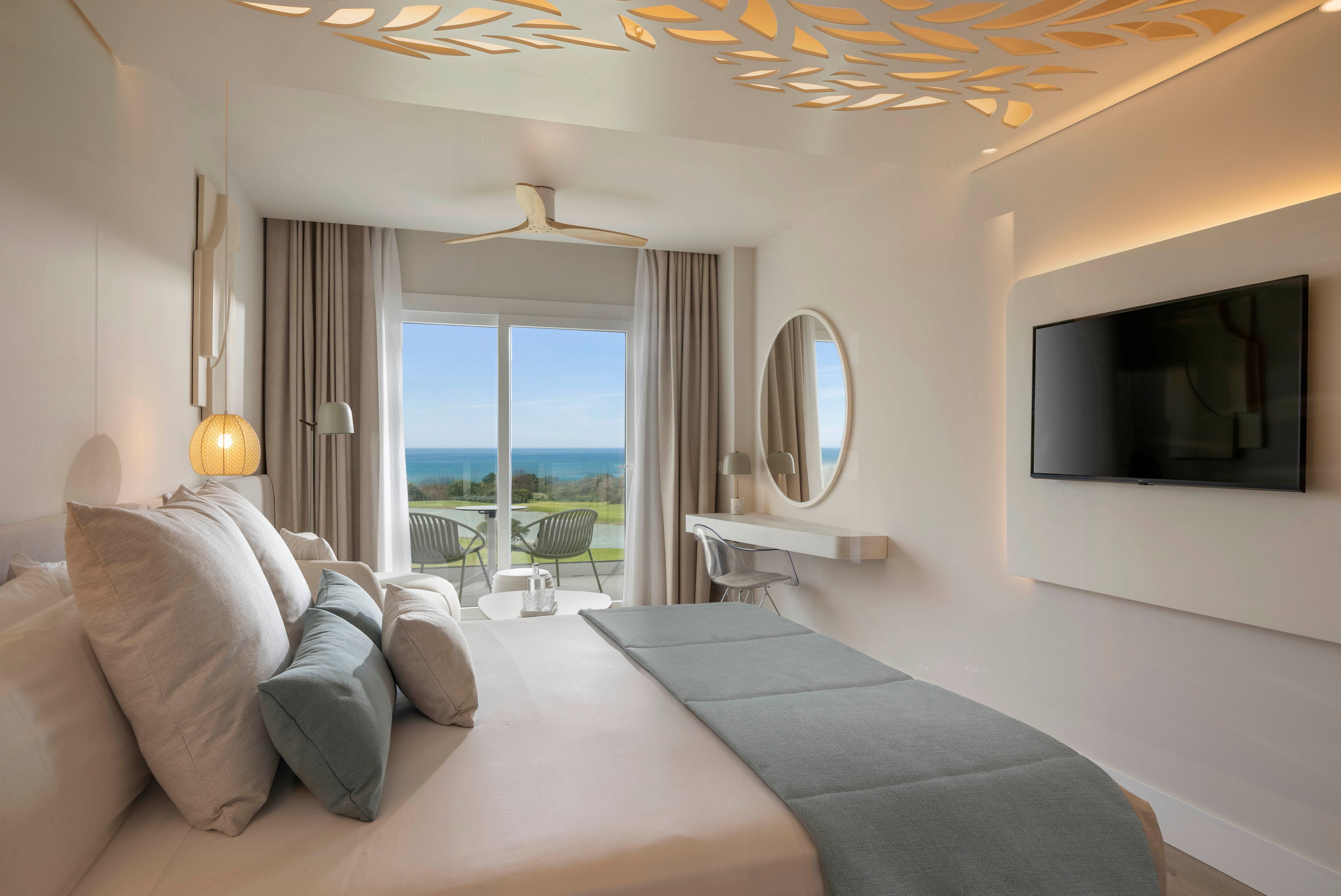 A contemporary hotel room features a neatly made bed with soft neutral linens and multiple pillows. Large glass doors open to a balcony with chairs, overlooking a green lawn and the distant ocean. The interior includes a wall-mounted flat-screen TV, a round mirror above a minimalist desk, and warm ambient lighting. Decorative cutouts on the ceiling and light-colored curtains contribute to the bright, airy atmosphere.