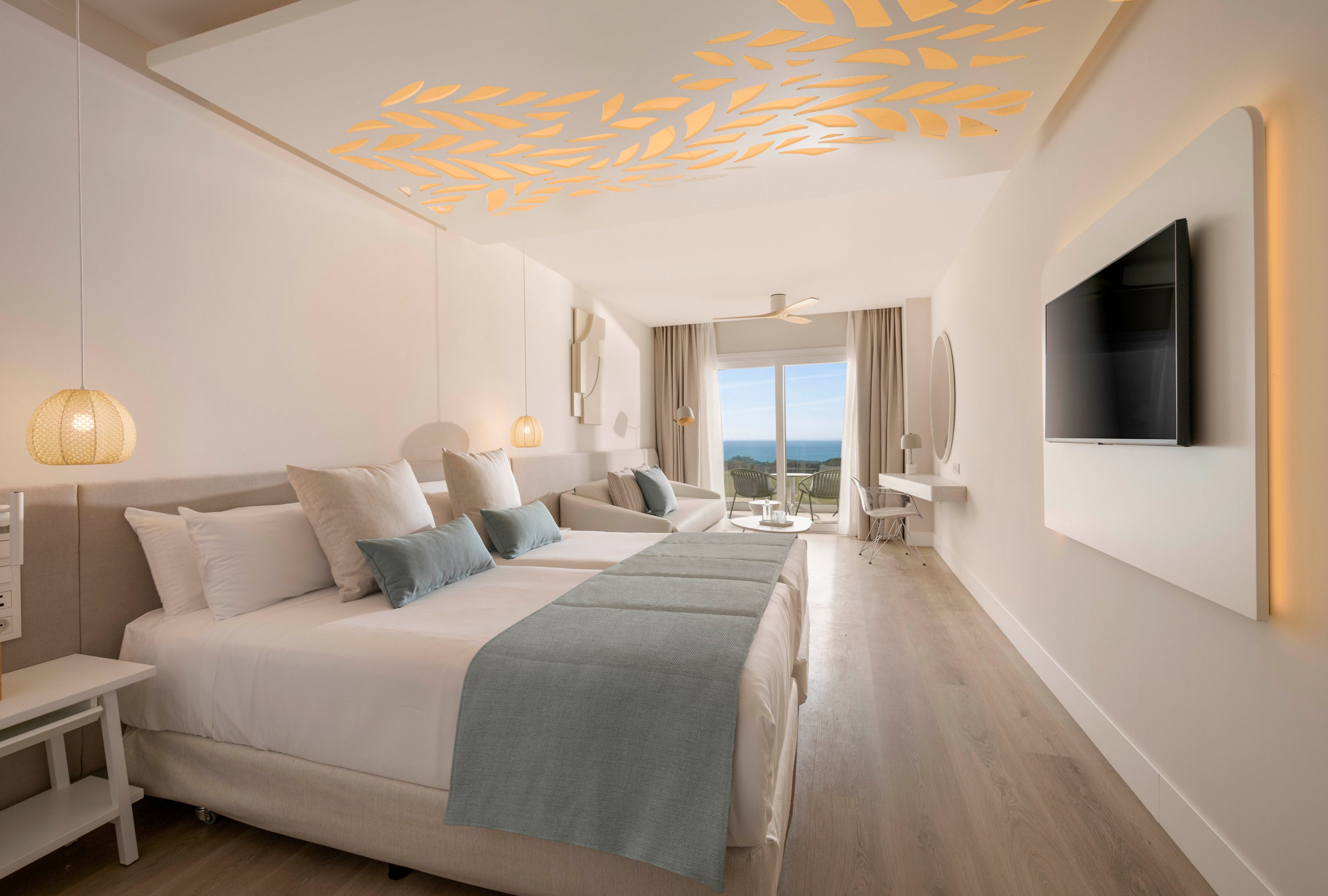A bright, modern hotel room features two neatly made beds with light blue throws and white linens. The space is decorated in a minimalist style with soft neutral tones and warm accent lighting in the ceiling and bedside lamps. A large flat-screen television is mounted on the wall opposite the beds, and a seating area with a sofa and desk extends toward a balcony. Floor-to-ceiling curtains frame a view of the ocean and clear sky outside, enhancing the airy, coastal atmosphere.