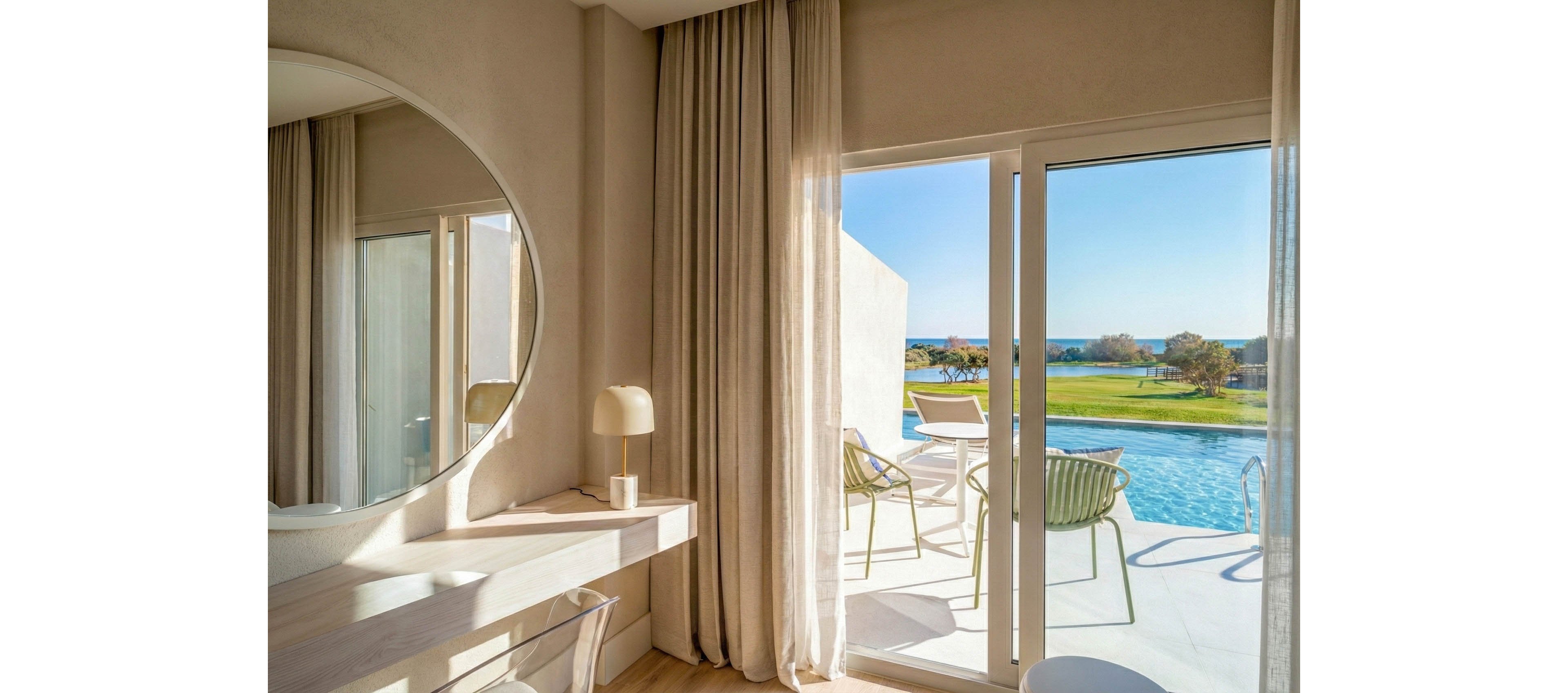 A bright, modern hotel room features a minimalist desk with a round mirror and a small table lamp. Sheer curtains frame large sliding glass doors that open onto a private terrace. Outside, lounge chairs and a small table sit beside a clear blue swimming pool. Beyond the pool, a manicured lawn and distant ocean create a serene coastal backdrop.