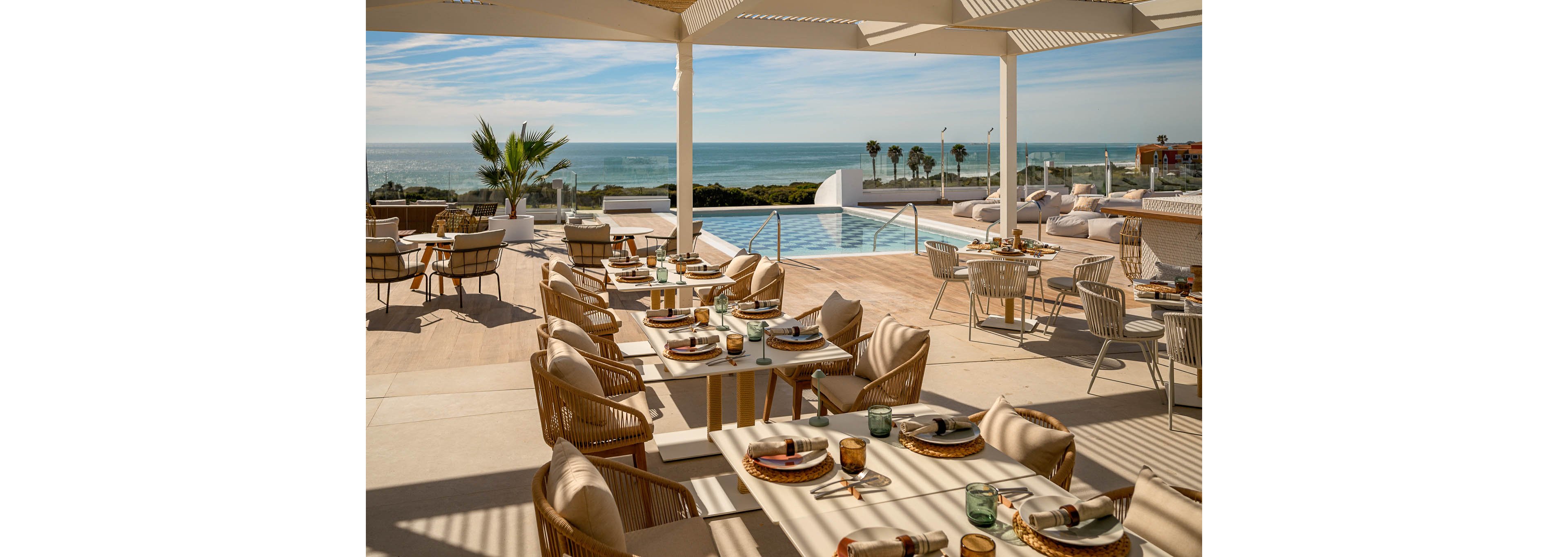 An elegant outdoor restaurant terrace overlooks a serene infinity pool and expansive ocean horizon. Sunlit dining tables are set with plates, glasses, and cutlery, ready for guests. Neutral-toned wicker chairs and loungers create a relaxed yet upscale atmosphere. Palm trees, clear skies, and calm water emphasize a coastal resort setting ideal for leisure and dining.