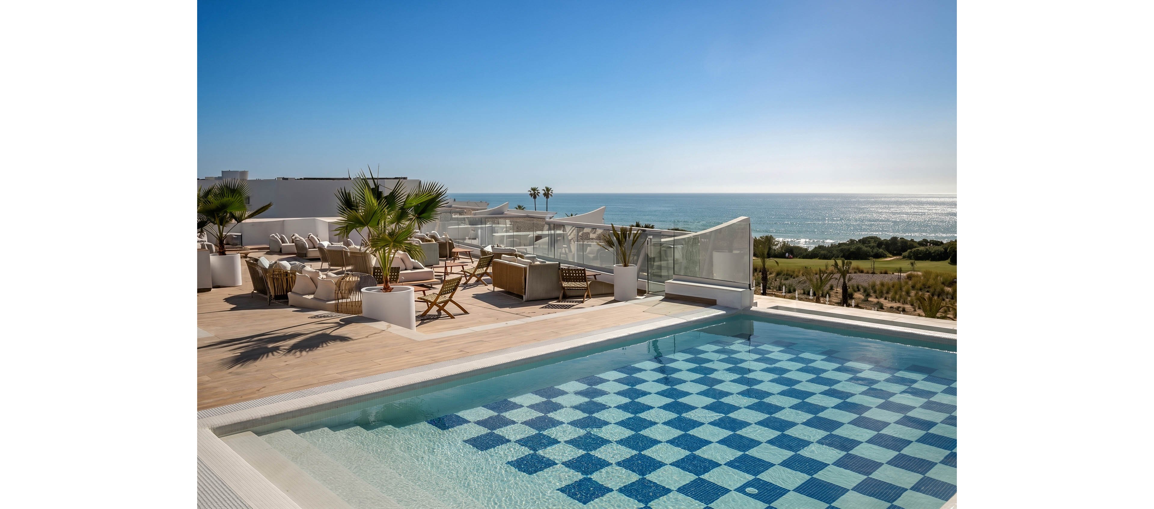 A modern rooftop swimming pool with a blue and white checkered tile pattern overlooks a vast ocean horizon. The spacious wooden deck is furnished with lounge chairs, sofas, and low tables arranged for relaxation and socializing. Palm trees in large planters add a tropical touch to the contemporary white architecture. Clear skies and bright sunlight create a serene, upscale resort atmosphere by the sea.