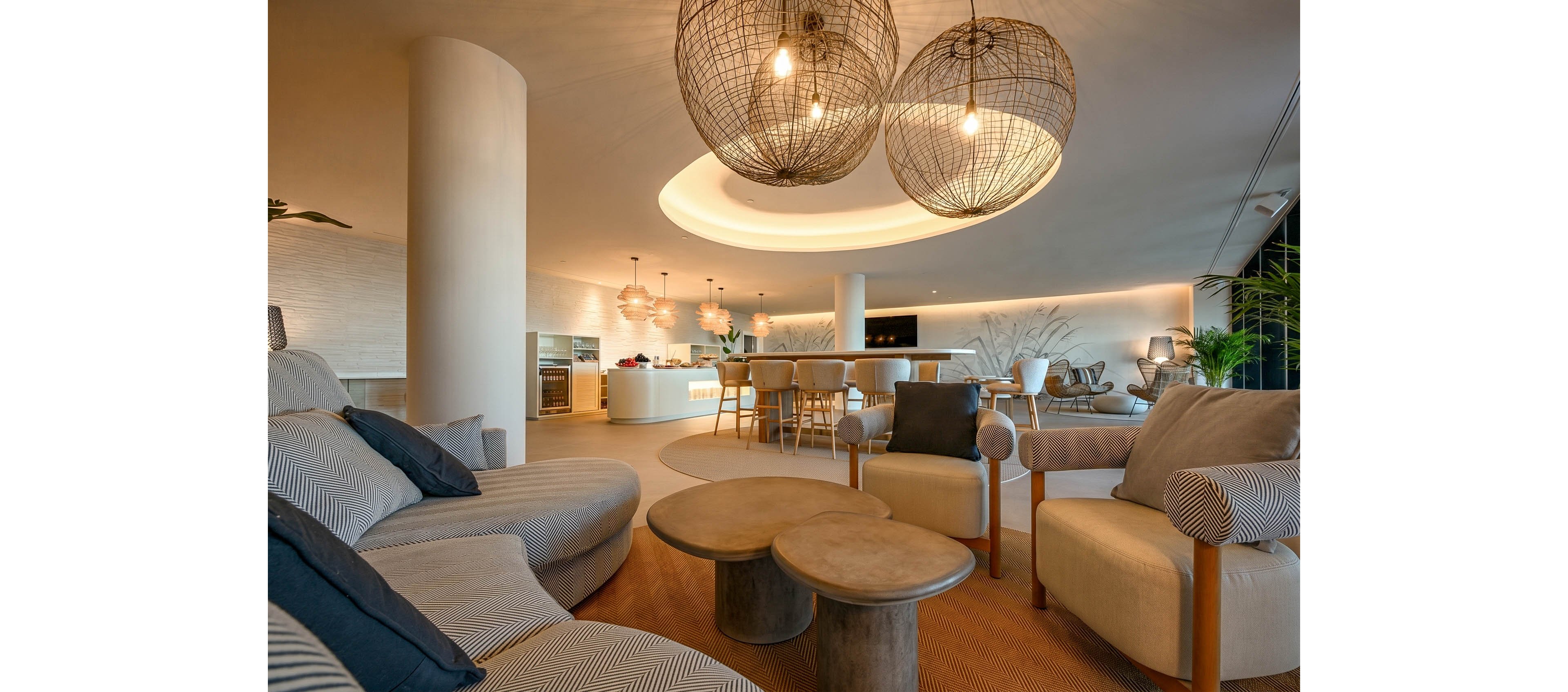 A contemporary lounge area features plush sofas and armchairs arranged around nested circular coffee tables. In the background, a sleek bar or kitchen counter with stools sits beneath a large recessed ceiling light. Oversized woven pendant lamps hang from the ceiling, casting a warm, inviting glow across the space. Neutral tones of beige, brown, and gray create a calm, sophisticated atmosphere with subtle decorative accents.