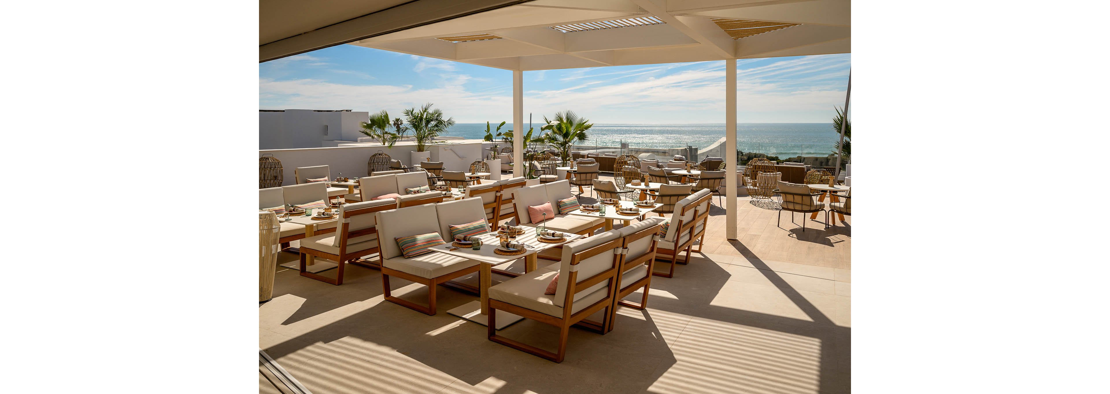 An open-air rooftop terrace features modern wooden lounge seating with cream cushions and colorful striped pillows. Low tables are set for casual dining, surrounded by woven chairs and potted plants. The space is covered by a partial pergola roof that lets in warm sunlight. In the background, the ocean and blue sky create a relaxed coastal atmosphere.