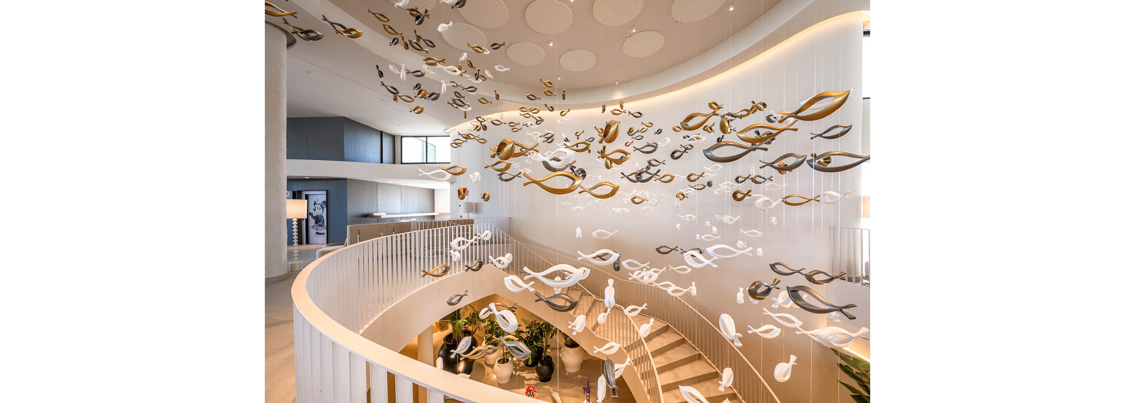 A contemporary interior lobby features a dramatic hanging sculpture composed of numerous abstract fish-like forms suspended from the ceiling. The artwork floats above a curved staircase with light-colored steps and sleek railings. Soft, indirect lighting highlights the white and metallic tones of the installation and the smooth architectural lines. Plants and seating areas below add warmth and texture to the sophisticated space.