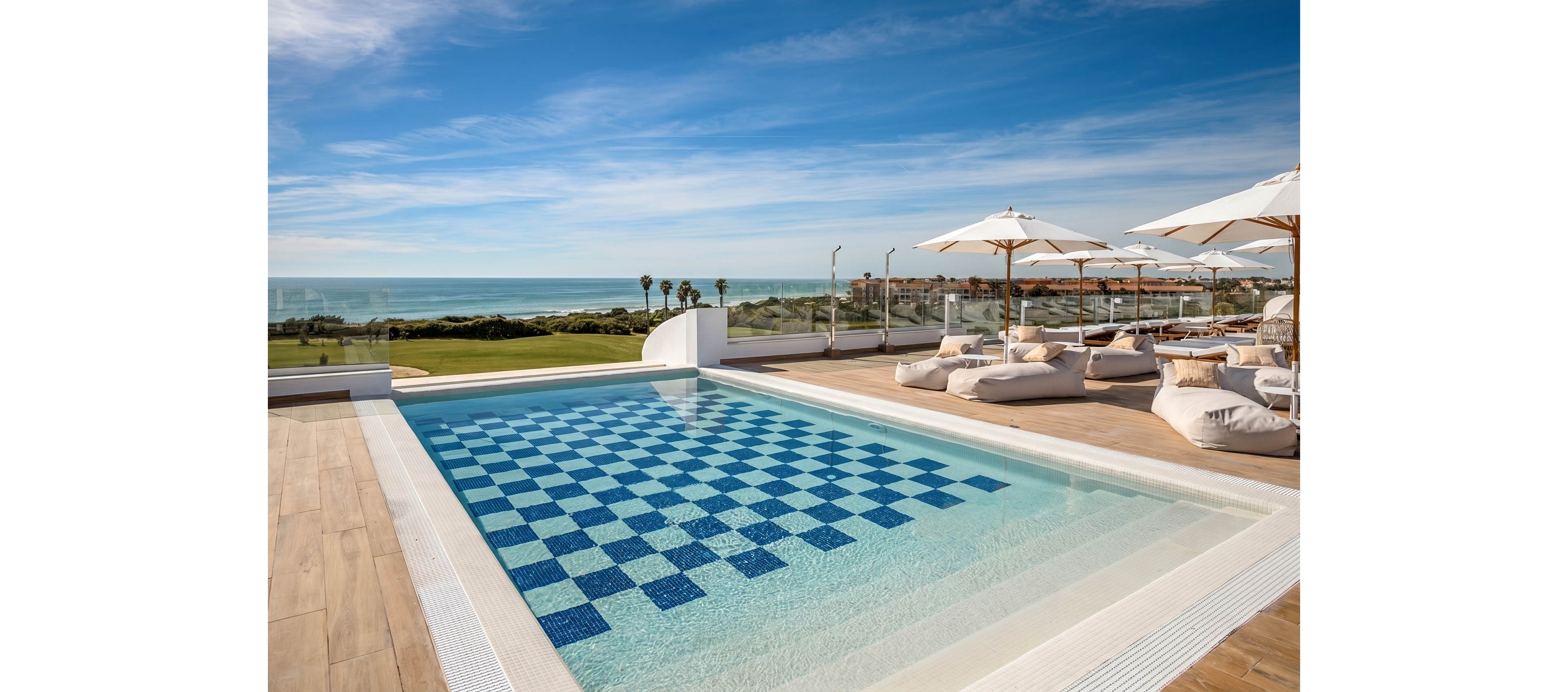 An elevated swimming pool with a blue and white checkerboard tile pattern sits on a wooden deck. White loungers and umbrellas line the terrace, creating a relaxed resort atmosphere. Glass railings reveal a scenic view of a green golf course and the ocean beyond. The bright blue sky and scattered clouds enhance the sunny, vacation-like mood.