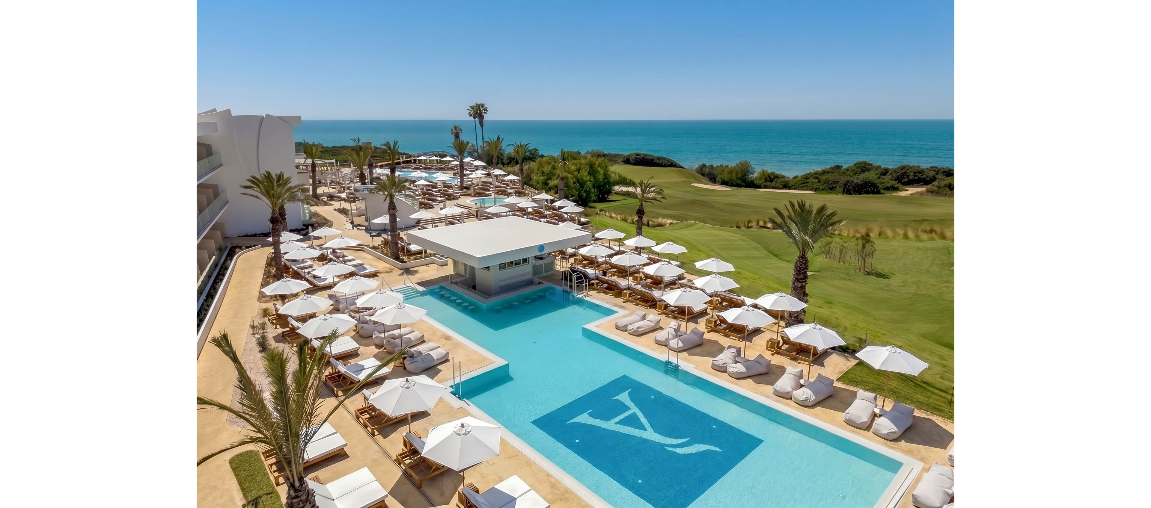 A luxurious resort pool area overlooks a scenic coastline and ocean under a clear blue sky. Numerous white sun loungers and umbrellas surround the turquoise swimming pool, creating an inviting relaxation space. Palm trees and manicured lawns add a tropical, upscale atmosphere. A modern pool bar sits at the center, while a golf course and natural greenery stretch toward the sea in the background.