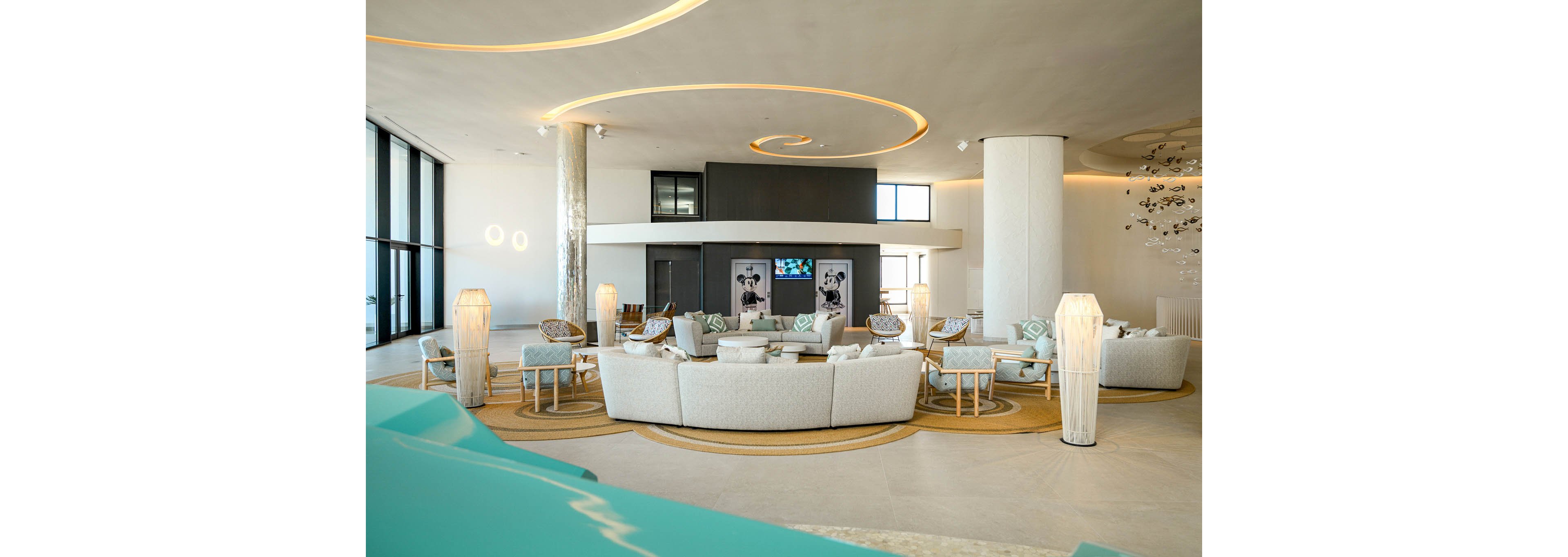 Spacious contemporary hotel lobby featuring multiple white sofas arranged in circular lounge areas. Soft neutral tones are accented by turquoise details and warm wooden side tables. Curved ceiling lights and tall floor lamps create an elegant, ambient glow. Large windows bring in natural light, highlighting the modern decor and polished flooring.