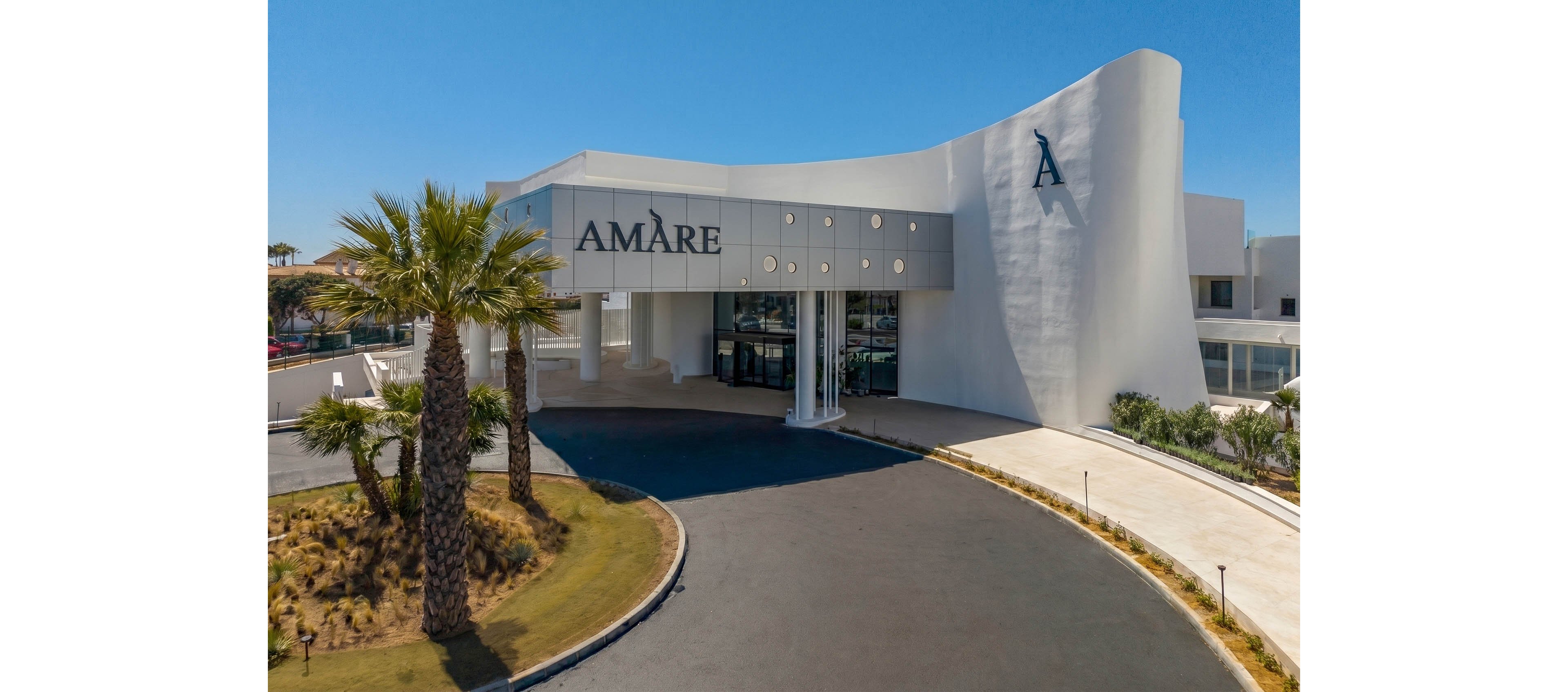 A contemporary white building labeled Amàre stands under a clear blue sky. The structure features curved walls, large glass entrance doors, and a covered drop-off area. A wide asphalt driveway curves toward the entrance, bordered by a manicured lawn and landscaped plants. Tall palm trees and low shrubs frame the scene, suggesting a warm, coastal or resort-like environment.