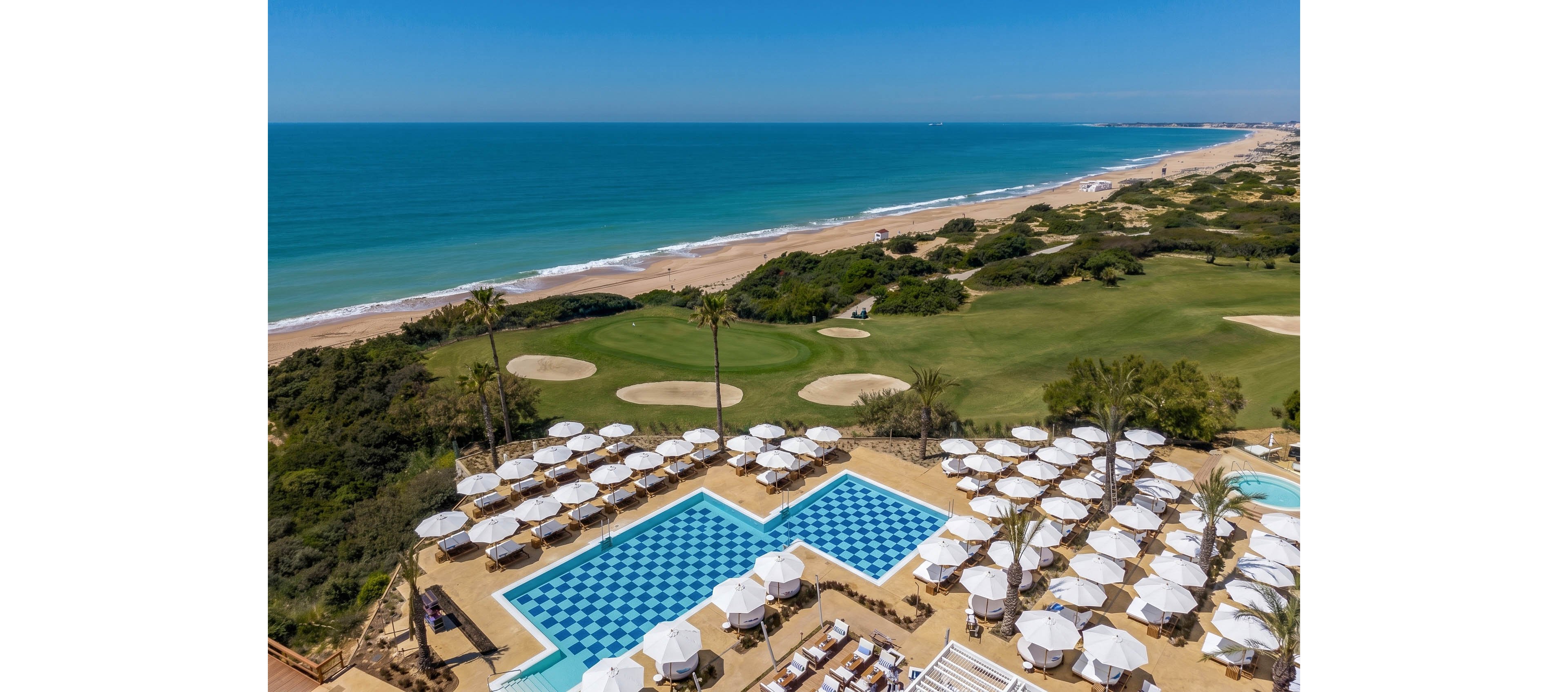 A coastal resort features a large outdoor pool area filled with white sun umbrellas and lounge chairs. The main swimming pool has a distinctive blue and white checkerboard pattern. Beyond the pool, a manicured golf course stretches toward a long sandy beach. Calm blue ocean water meets a clear sky, creating a bright and relaxing vacation atmosphere.