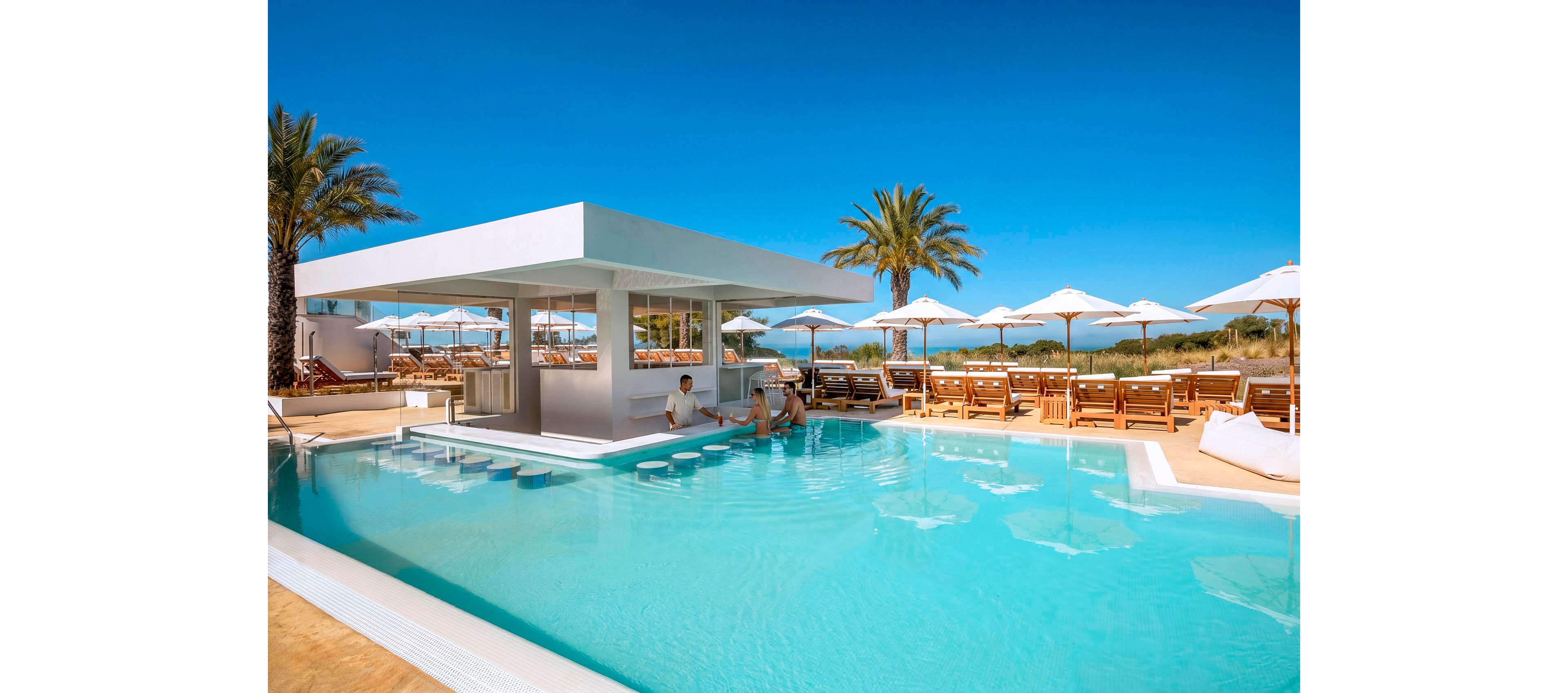An upscale outdoor swimming pool features a modern white swim-up bar at its center. Guests relax on submerged bar stools and nearby loungers under white umbrellas. Palm trees and clear blue skies create a tropical vacation atmosphere. The scene suggests a sunny, tranquil day at a beachside resort.