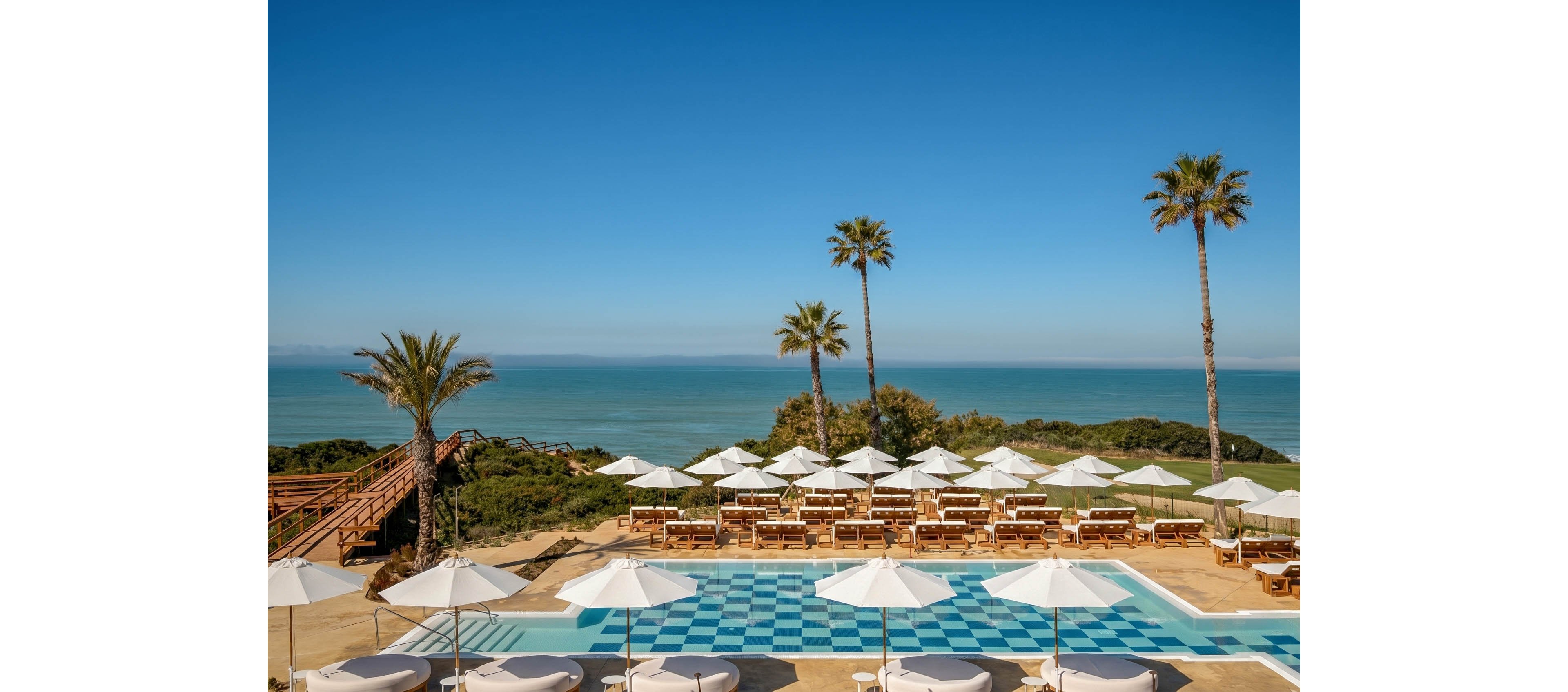 An oceanfront resort pool area overlooks a calm, expansive sea under a clear blue sky. White umbrellas and wooden sun loungers are neatly arranged around a checkered blue-and-white swimming pool. Tall palm trees and lush greenery frame the scene, with a wooden boardwalk leading toward the beach. The bright sunlight and vivid colors create a relaxed, luxurious vacation atmosphere.