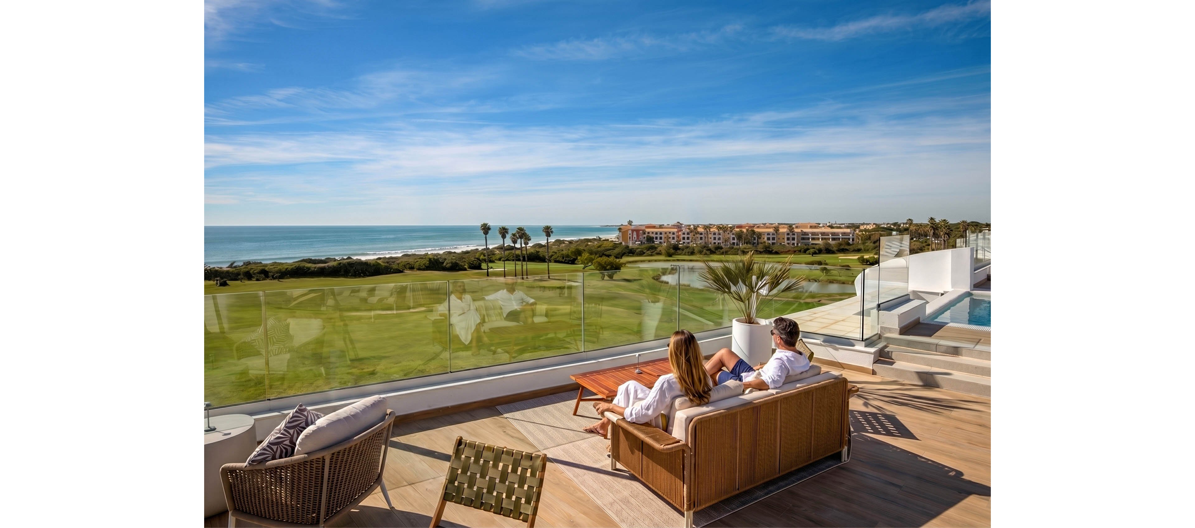 A couple lounges on a modern wooden sofa on a spacious terrace overlooking a golf course and the ocean. The setting features glass railings, a swimming pool, and contemporary outdoor furniture under a clear blue sky. In the distance, low-rise resort buildings and palm trees line the coastline. The overall scene conveys leisure, comfort, and upscale coastal living.