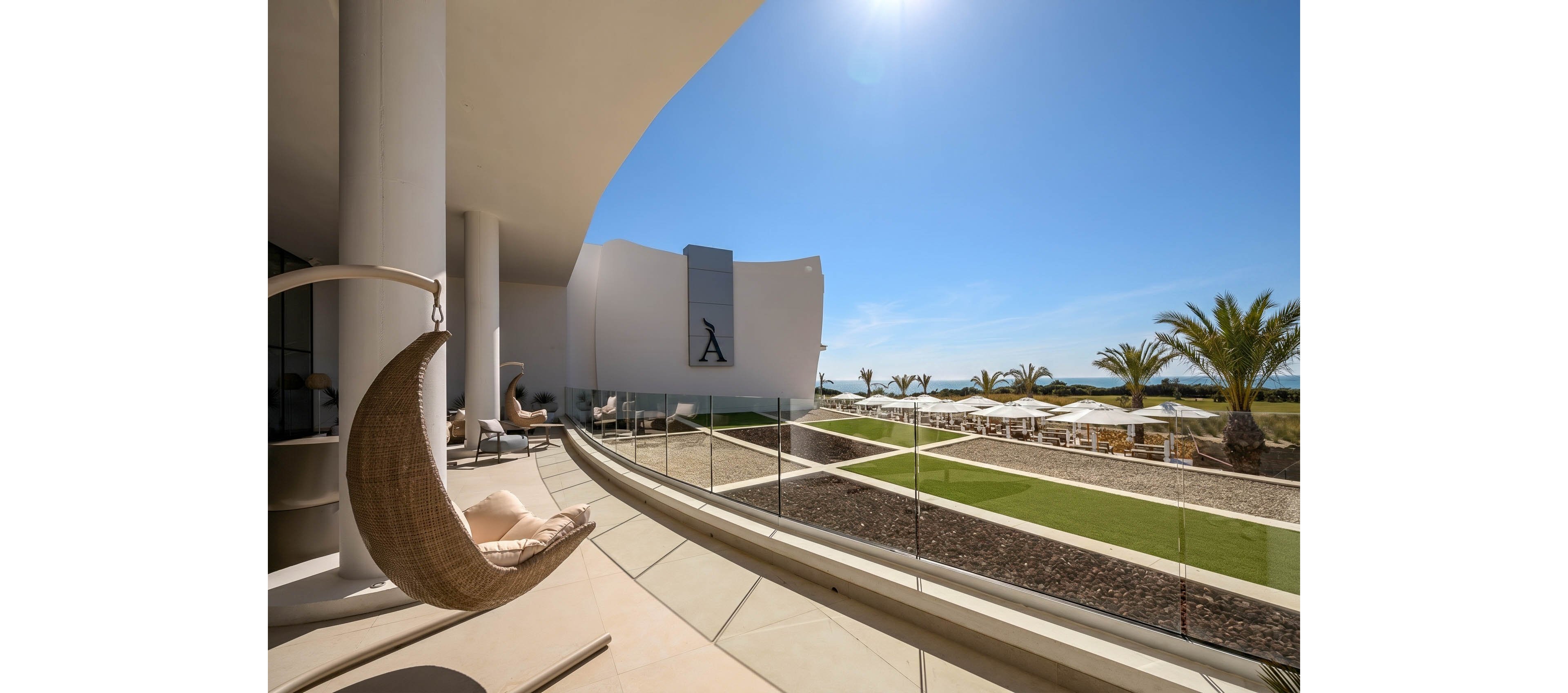 A contemporary resort terrace overlooks a manicured lawn and distant ocean under a clear blue sky. The curved balcony features glass railings and a hanging wicker chair in the foreground. In the background, palm trees and rows of white sun loungers with umbrellas line the outdoor relaxation area. Bright sunlight and clean architectural lines create a luxurious, tranquil coastal atmosphere.