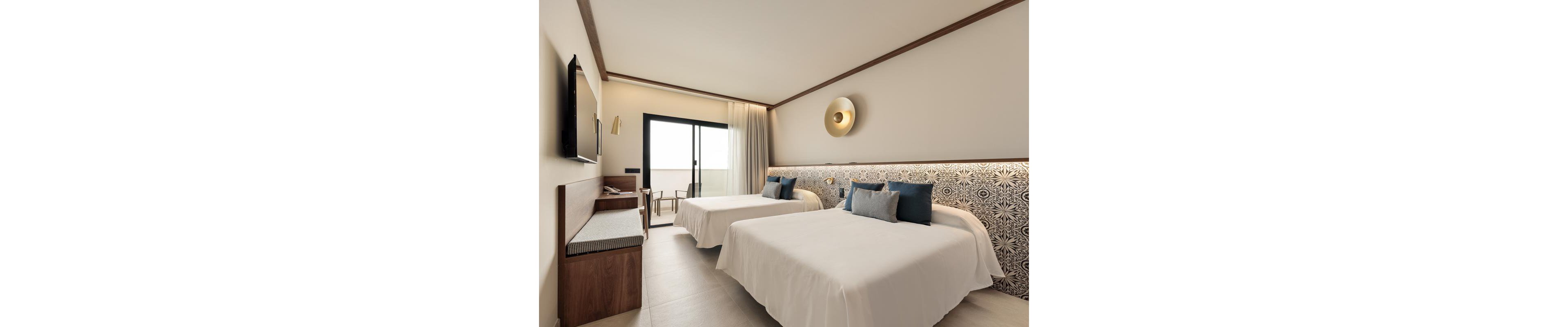 The image shows a contemporary hotel room featuring two double beds with patterned headboards and accent pillows. The space is well-lit, with neutral tones and minimalist decor, including a wall-mounted TV, desk, and a stylish wall lamp. A sliding glass door leads to a balcony, allowing natural light to fill the room. The overall mood is calm and inviting, ideal for relaxation or leisure travel.