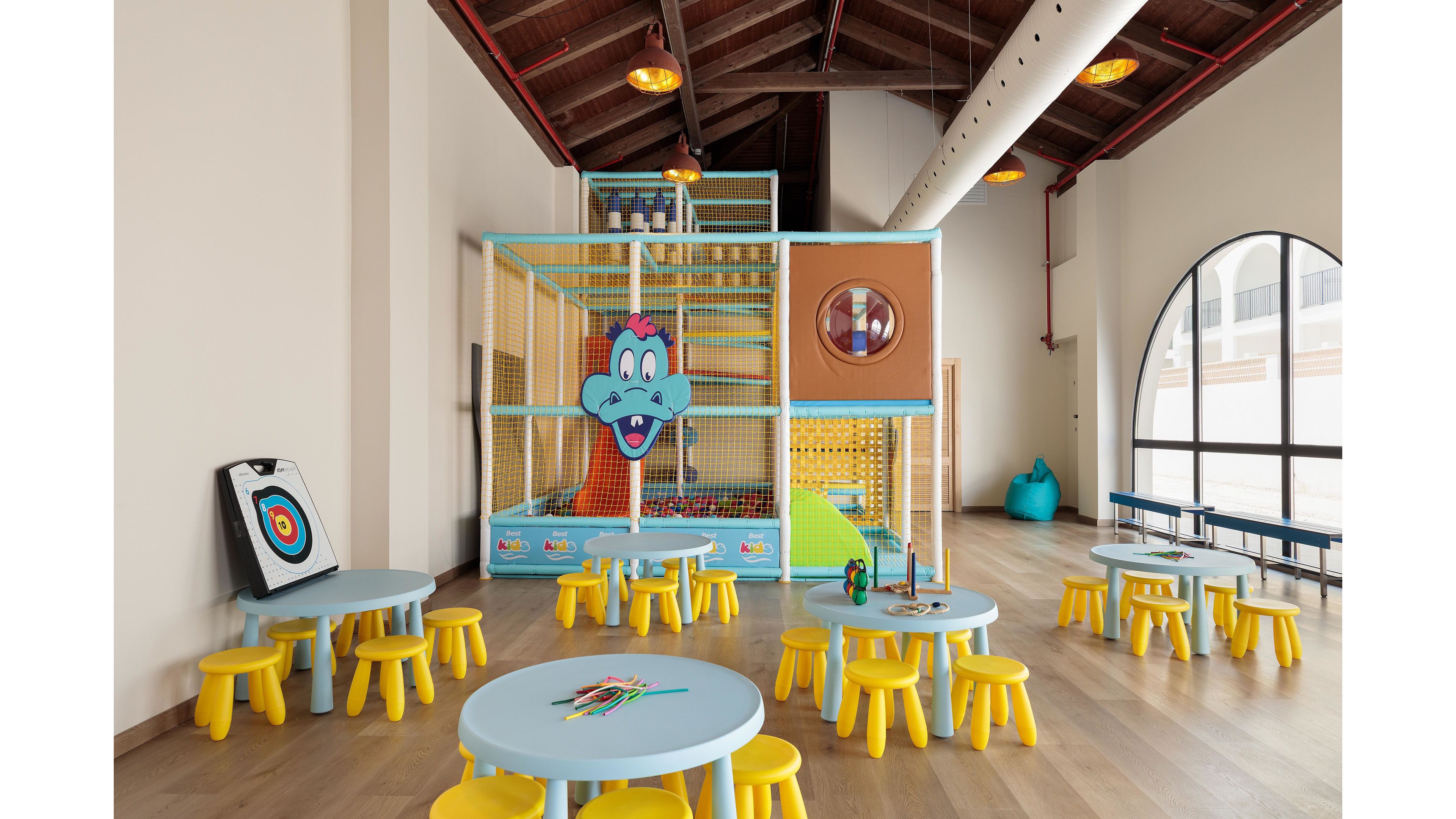 A spacious indoor playroom features a large climbing structure with a cartoon animal face and a ball pit. The room is furnished with small round tables and yellow stools, designed for young children. The setting is bright and airy, with wooden floors, high ceilings, and large arched windows letting in natural light. A target board and various toys are visible, creating a playful and inviting atmosphere.