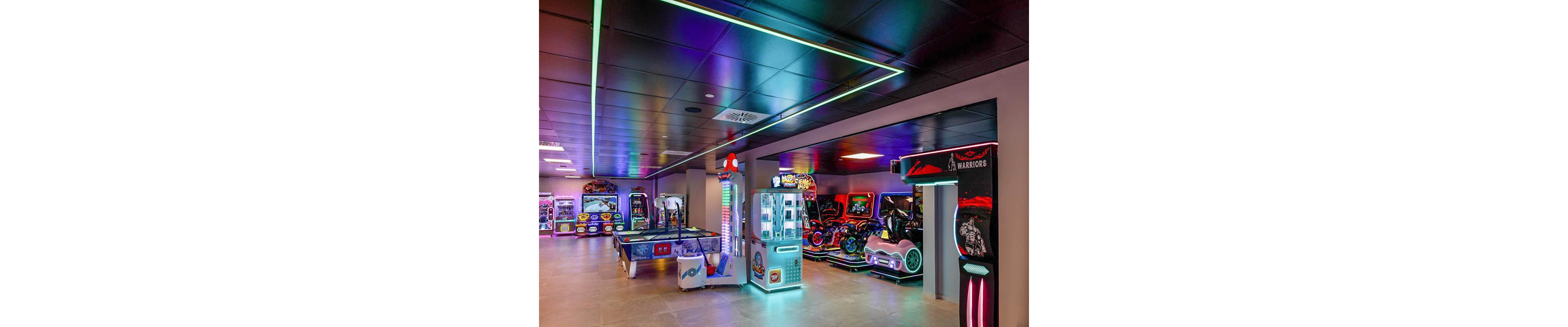A vibrant arcade room features a variety of electronic game machines, including racing simulators, air hockey, and claw machines. The setting is indoors with colorful neon lighting reflecting off the ceiling and floor, creating a lively and energetic atmosphere. Several machines display bright screens and illuminated controls, enhancing the playful mood. No visible people are present, and no explicit numeric values or unmistakable brand names are seen.