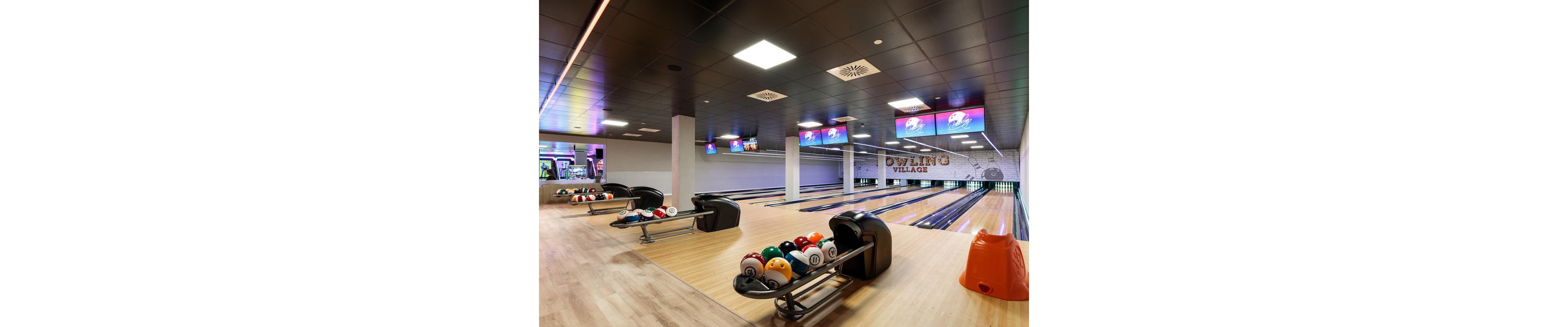 The image shows a contemporary bowling alley featuring multiple lanes and racks of vibrant bowling balls. The setting is well-lit with overhead lights and several digital screens displaying graphics. The wall in the background has visible text reading 'Bowling Village,' adding to the venue's identity. No people are present, and the overall mood is clean and inviting.