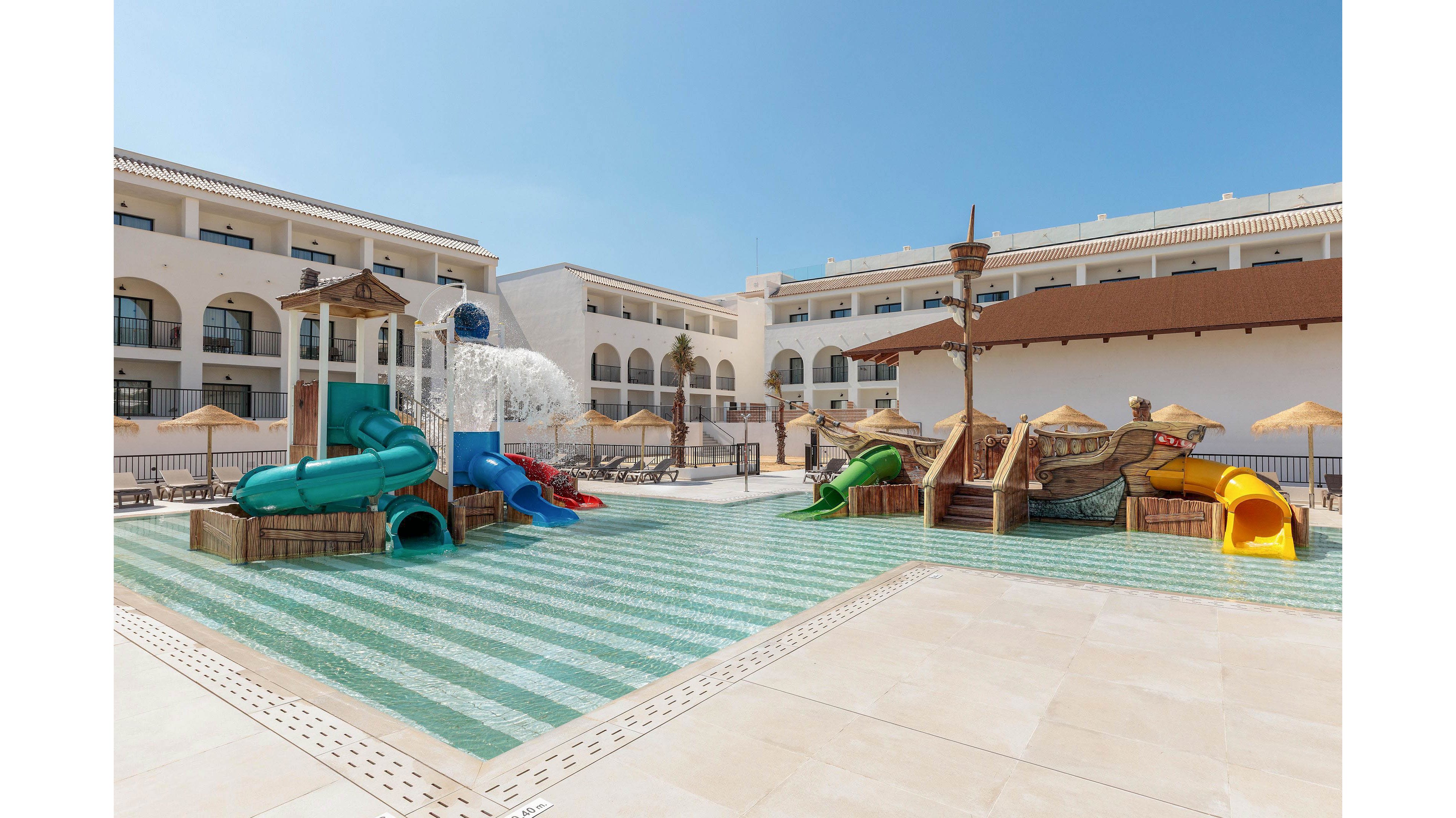 The image shows a children's water play zone featuring colorful slides and splash structures, set within a hotel courtyard. The area is surrounded by sun loungers and umbrellas, with the hotel building visible in the background. The setting is bright and sunny, evoking a cheerful and family-friendly mood. No visible text or numeric values are present in the image.