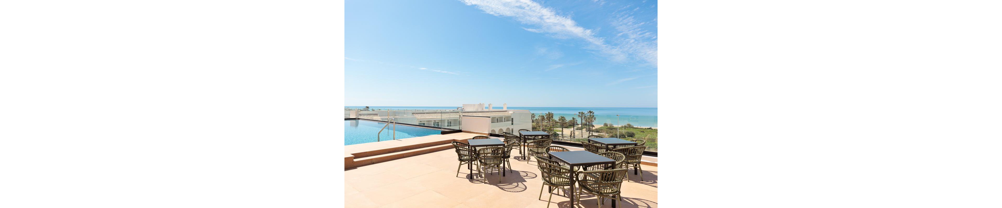 The main subject is an outdoor terrace featuring several black tables and chairs arranged for dining. The setting includes a clear view of a swimming pool and the ocean, with palm trees and modern white buildings in the background. The visuals highlight a bright, sunny day with blue skies and minimal clouds, creating a relaxed and inviting atmosphere. No visible text or numeric values are present in the image.