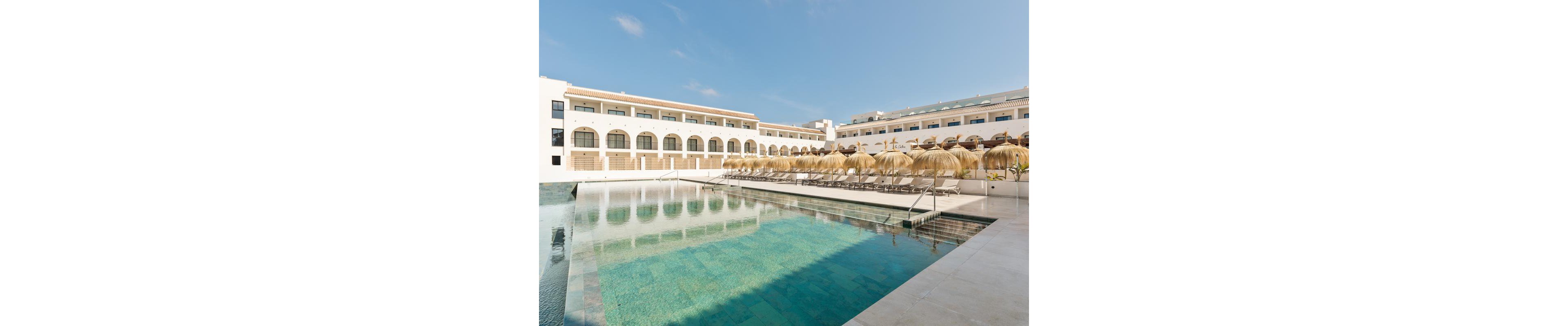 The image features a contemporary hotel complex with multiple stories and arched windows, set around a large outdoor swimming pool. Numerous sun loungers and straw parasols line the poolside, creating a relaxing vacation atmosphere. The clear blue sky and tranquil water reflect the clean architectural lines and inviting leisure setting. No visible text or numeric values are present in the image.