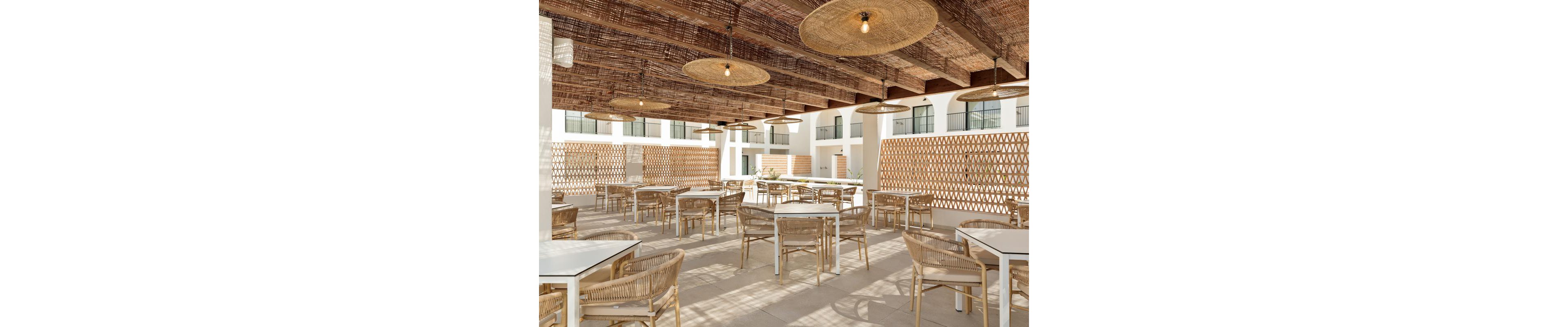 Spacious open-air dining area featuring white square tables and wicker chairs arranged under a wooden pergola. The ceiling is adorned with woven rattan light fixtures, creating a warm and inviting atmosphere. Natural light fills the space, highlighting the geometric lattice walls and neutral color palette. No people are present, emphasizing the tranquil and relaxed setting.