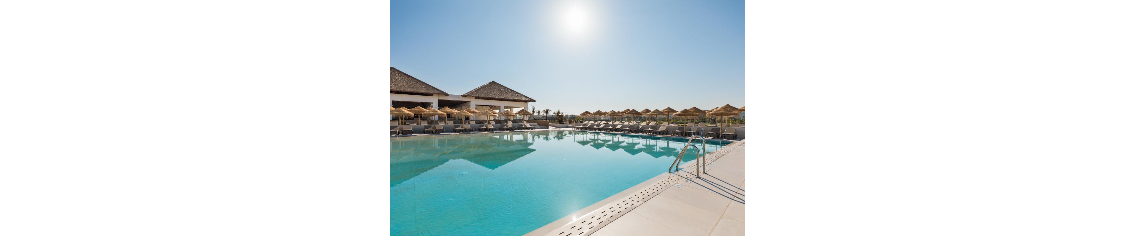 A large outdoor swimming pool is featured in a resort setting under a clear blue sky. Numerous lounge chairs and straw umbrellas line the poolside, creating a relaxing atmosphere. The sun is shining brightly, casting reflections on the water and highlighting the tranquil mood. No people are visible, emphasizing the serene and peaceful environment.