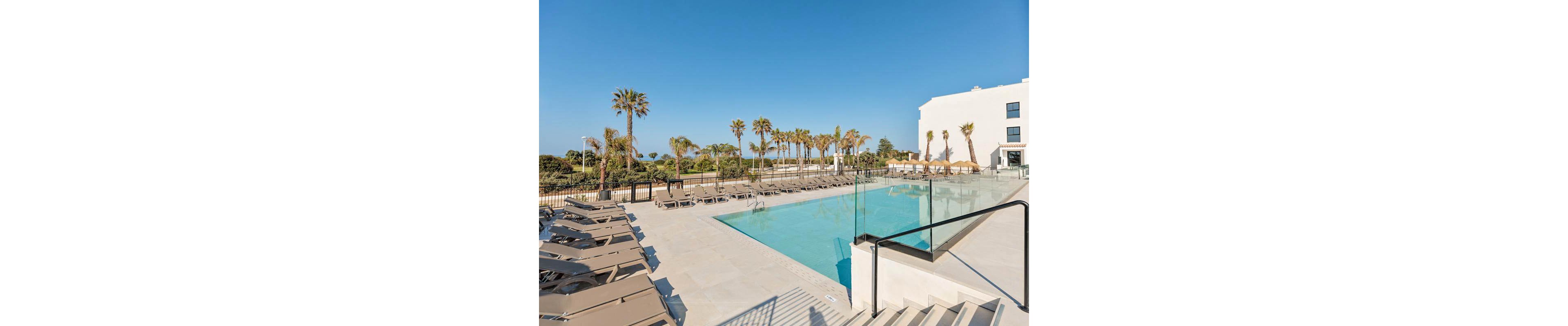 The image features a contemporary outdoor swimming pool surrounded by numerous beige loungers and palm trees. The setting is a sunny, clear day with a blue sky, and the pool area is adjacent to a white building. Glass railings and clean lines contribute to a minimalist, resort-like atmosphere, evoking relaxation and leisure. No visible text or numeric values are present in the image.