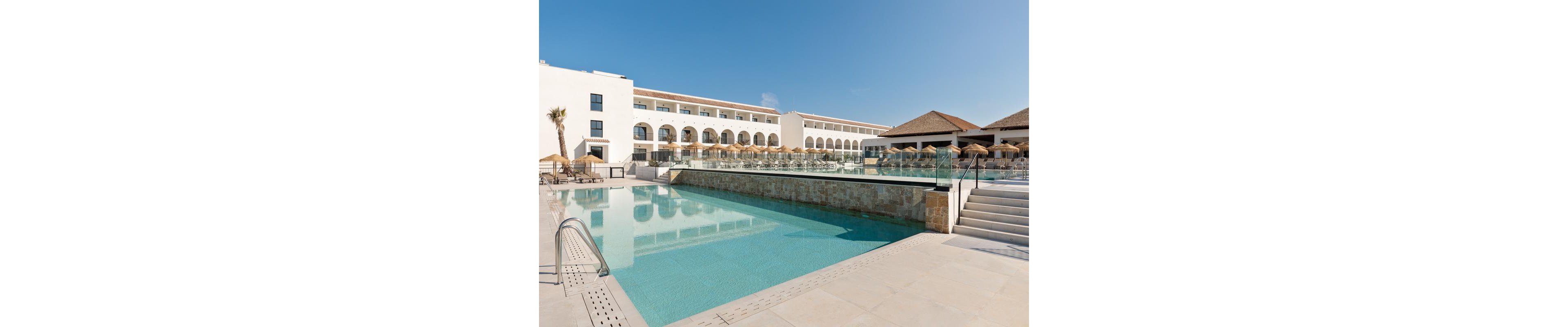 A contemporary hotel building features a large outdoor swimming pool surrounded by sun loungers and umbrellas. The setting is bright and sunny, with clear blue skies and clean architectural lines. The pool area is spacious, with glass railings and steps leading to a terrace. No visible text or numeric values are present in the image.