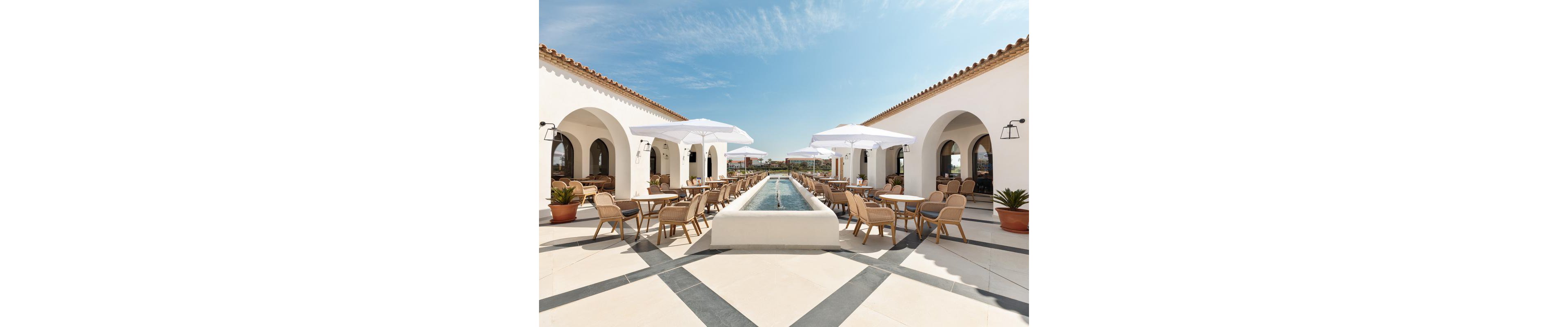 The image shows a spacious outdoor dining area featuring white Mediterranean-style architecture with arched doorways. Wooden chairs and tables are arranged around a central water feature, shaded by large white umbrellas. The setting is bright and sunny, with clear blue skies and potted plants adding to the relaxed ambiance. No visible text or numeric values are present.