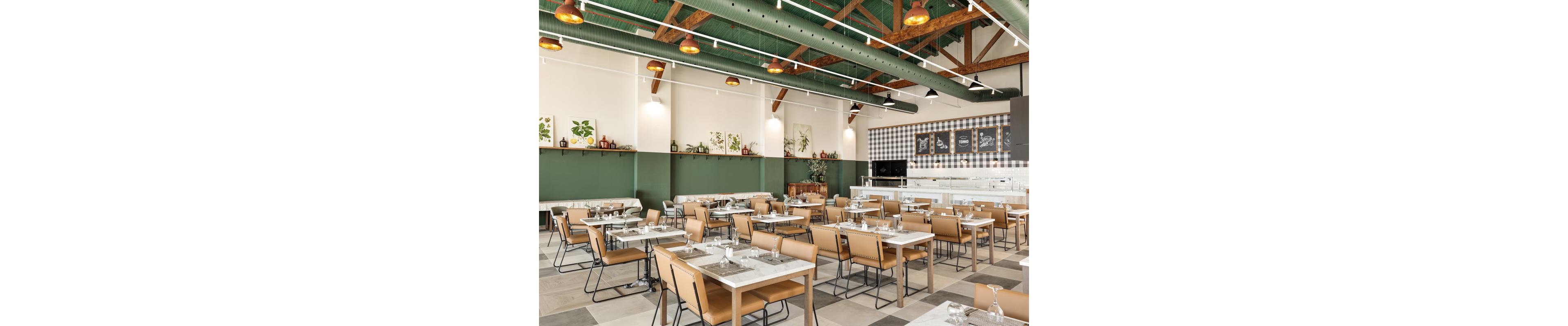 The image shows a spacious cafeteria with neatly arranged tables and tan chairs. The setting features green walls, wooden ceiling beams, and decorative plant artwork, creating a fresh and inviting atmosphere. The cafeteria is well-lit with pendant lights and has a clean, contemporary style. The counter area displays menu boards with visible chalk-style text, but no specific brands or numbers are clearly identifiable.