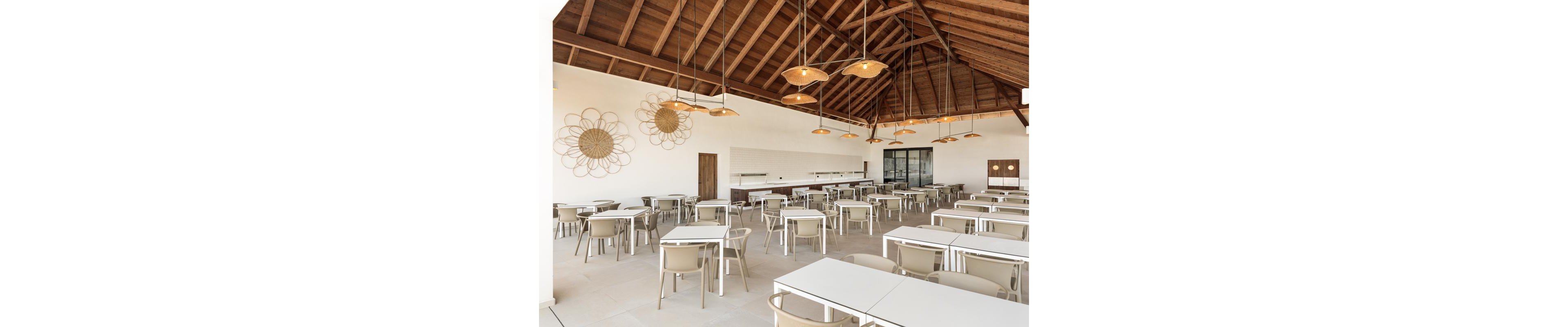 The image shows a spacious dining area featuring white tables and beige chairs arranged neatly under a high wooden ceiling. The setting is bright and airy, with decorative wall art and pendant lights adding visual interest. The space appears empty, emphasizing its clean and minimalist style. Large windows and doors provide natural light, enhancing the warm and inviting atmosphere.