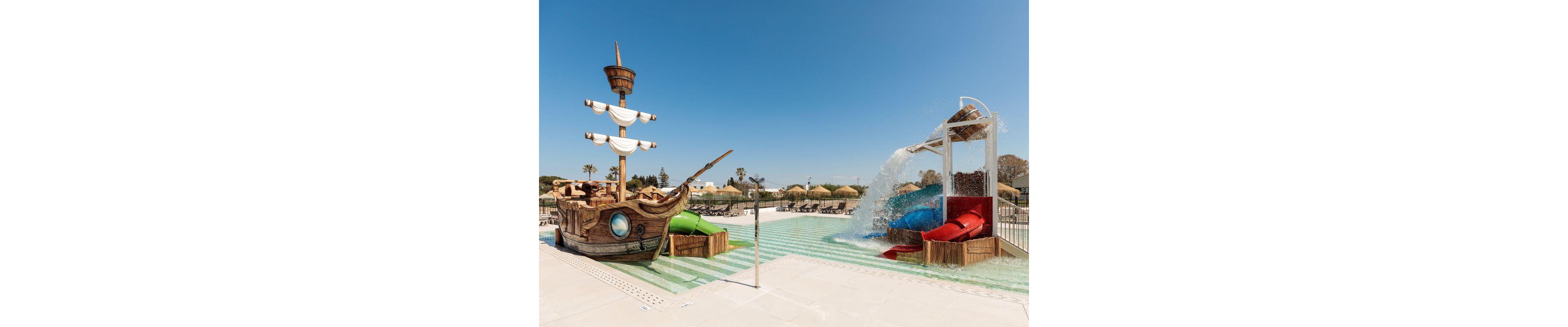 A vibrant outdoor water playground features a pirate ship structure and colorful slides. The setting is a sunny pool area with straw umbrellas and lounge chairs in the background. Water cascades over the slides, creating a playful atmosphere. The scene is designed for children, with bright colors and whimsical elements. No visible text or numeric values are present in the image.