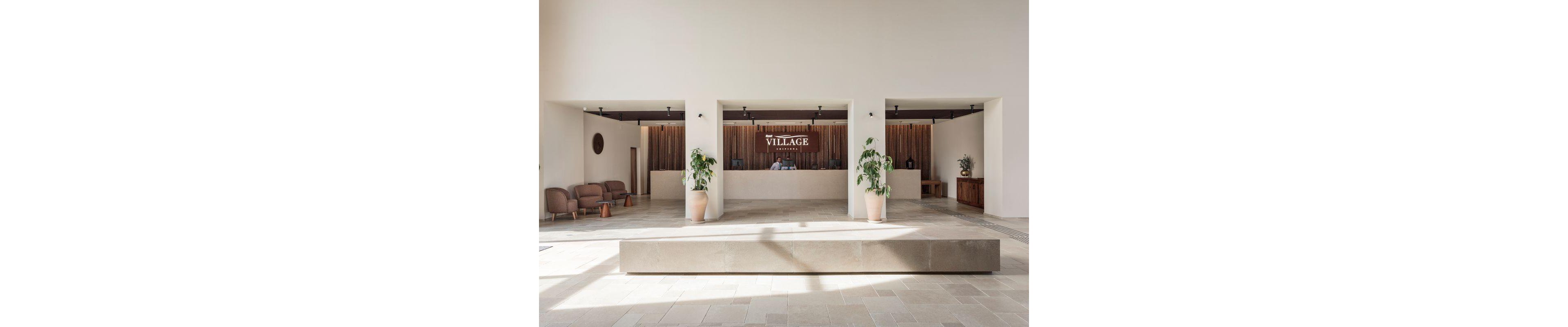 The image shows a spacious hotel reception area with three counters and a prominent 'Village' sign behind the central desk. The setting features minimalist decor, neutral tones, and large potted plants, creating a welcoming atmosphere. Two people are visible at the main counter, and comfortable seating is arranged to the left. The overall mood is calm and contemporary, with natural light enhancing the space.