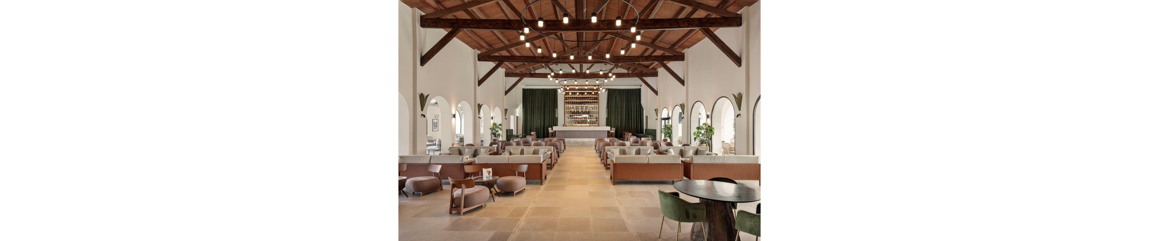 The image features a large, open lounge area with a central bar at the far end. The space is furnished with contemporary sofas, armchairs, and tables arranged in symmetrical rows. High wooden ceilings with exposed beams and stylish lighting fixtures create a warm, inviting atmosphere. Arched windows and green plants add to the airy, elegant setting, while neutral tones dominate the palette.