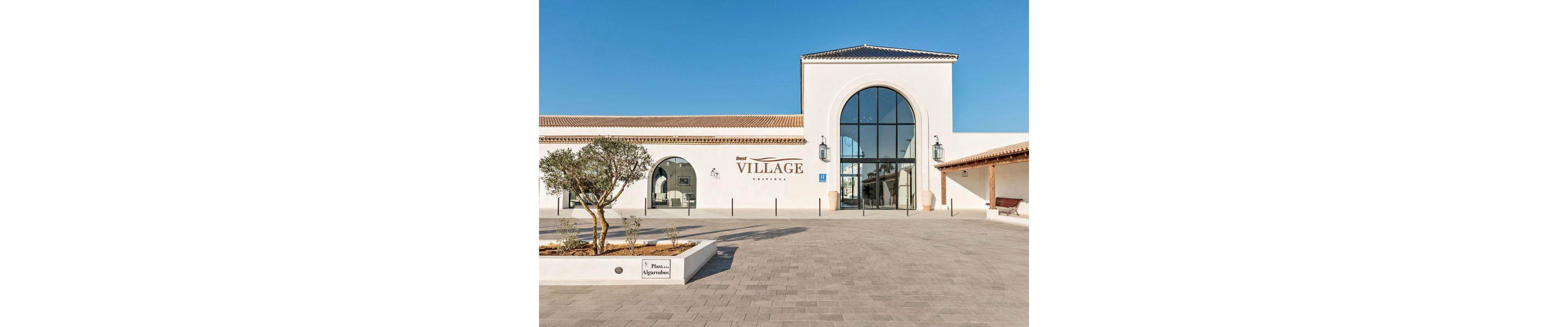 The image shows the entrance to a market named 'VILLAGE MARKET,' featuring a large glass archway and white stucco walls. The setting is an open plaza with a paved courtyard, a small tree planter labeled 'Plaza Algarbina,' and clear blue skies. Architectural details include lantern-style wall lights and a bench under a covered walkway. Visible text includes 'VILLAGE MARKET' and 'Plaza Algarbina.'
