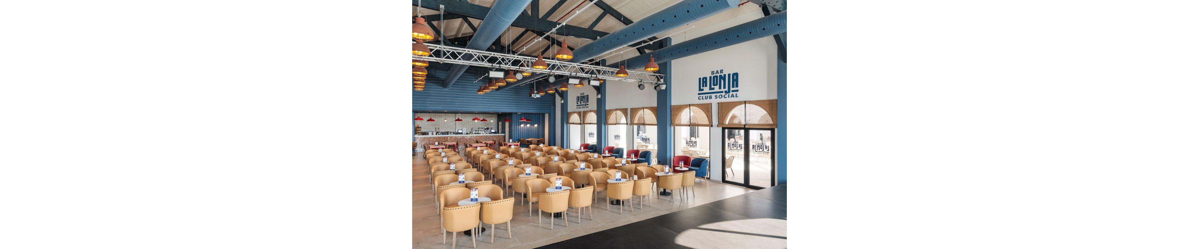 The image shows the interior of Bar La Lonja Club Social, featuring rows of tan and red chairs arranged around tables. The setting is a modern, airy bar with blue structural beams, large windows, and ample natural light. The mood is inviting and social, with a clean palette of blue, tan, and red. Visible text includes 'BAR LA LONJA CLUB SOCIAL' on the wall and doors.