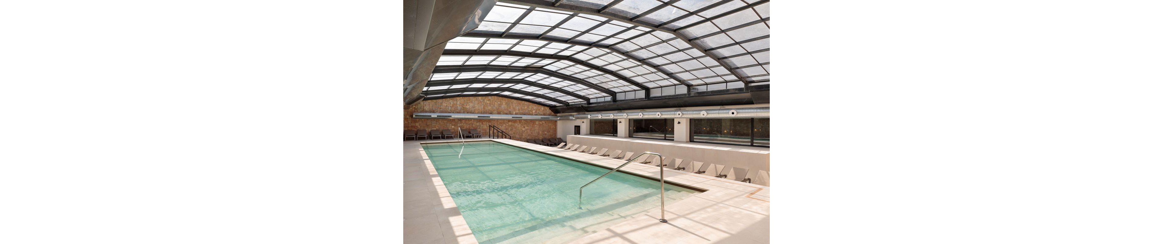 A spacious indoor swimming pool is featured beneath a large glass roof, allowing natural light to fill the area. The pool is surrounded by numerous lounge chairs and a sleek metal handrail, creating a relaxing atmosphere. The setting is clean and contemporary, with neutral tones and minimalist design elements. No visible text or numeric values are present in the image.