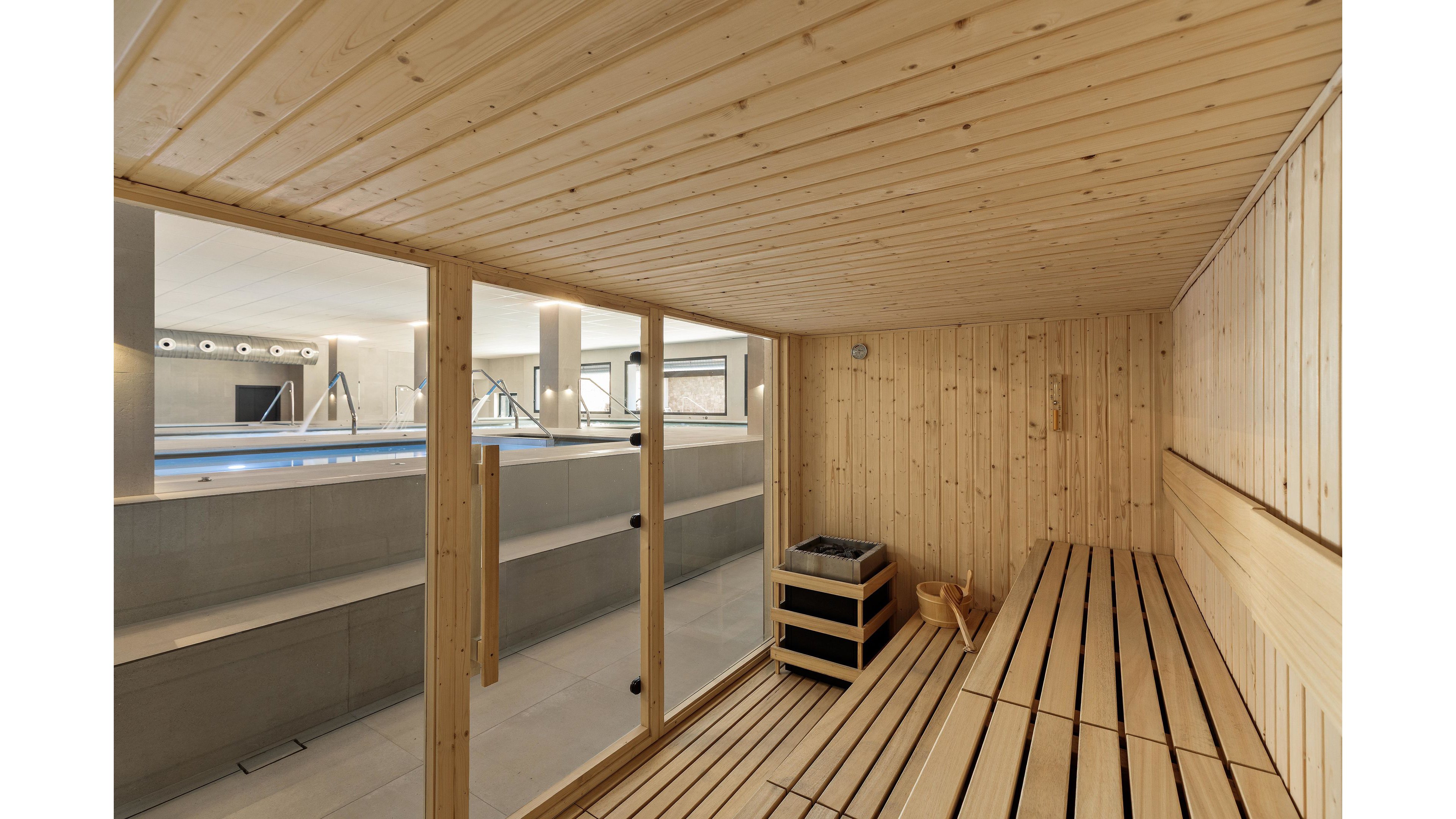 The main subject is a contemporary wooden sauna with clean lines and light wood paneling. The sauna features benches, a heater, and a bucket with ladle, all visible through a glass wall. Outside, an indoor swimming pool area with minimalist design and neutral tones can be seen. The setting is bright and spacious, evoking a calm and relaxing mood.