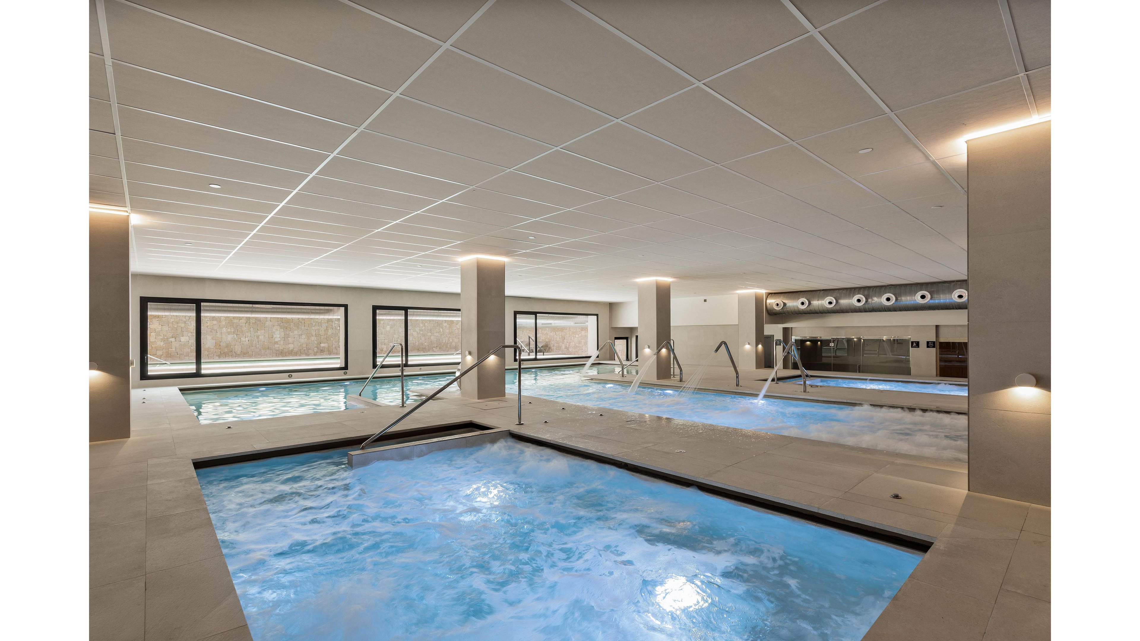 The image features a spacious indoor spa area with multiple pools, including a hot tub and lap pool. The setting is contemporary, with clean lines, neutral tones, and large windows allowing natural light. The water in the pools is illuminated with a soft blue glow, enhancing the tranquil mood. No people are visible, emphasizing a serene and peaceful environment.