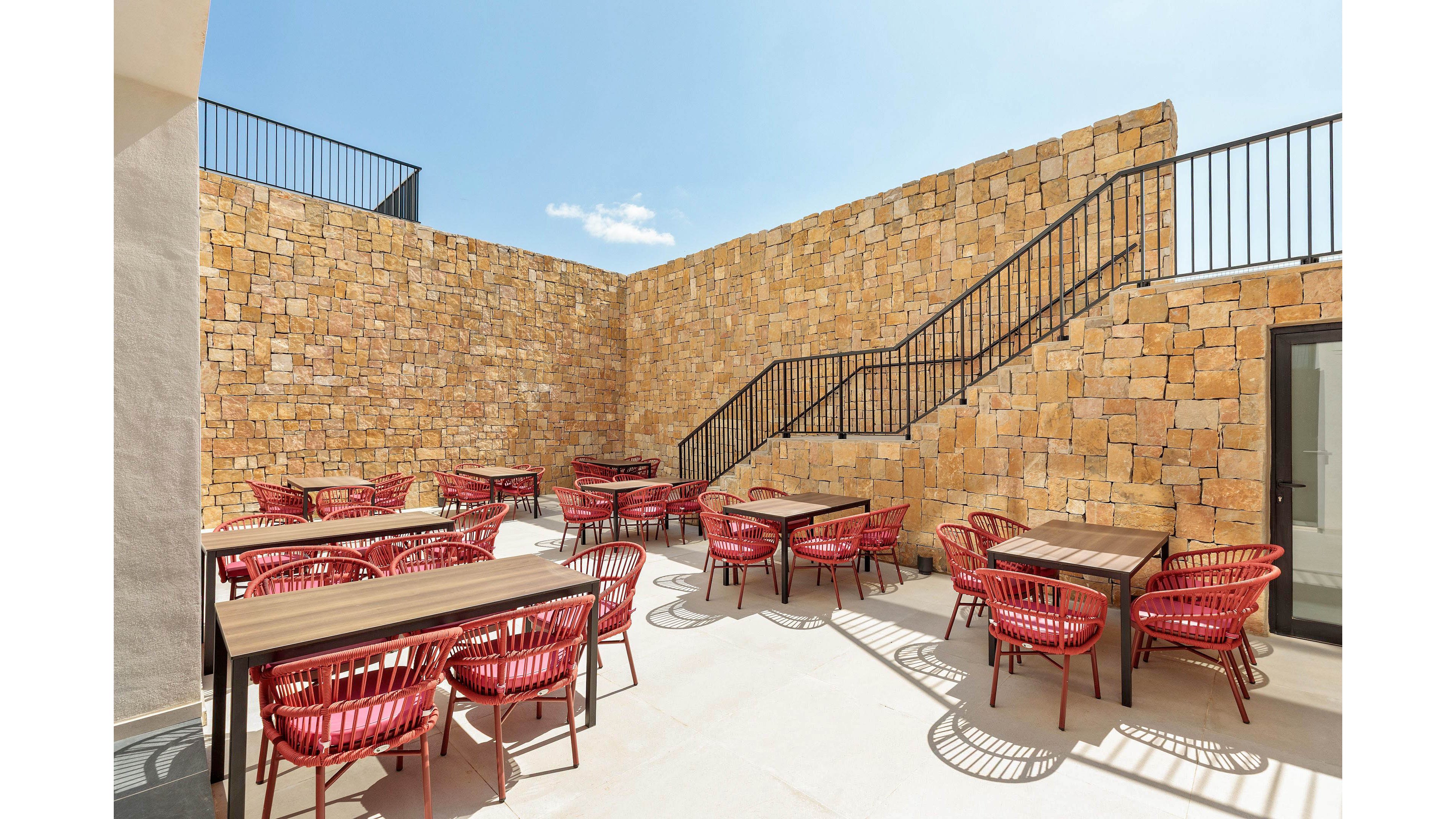 The image shows an outdoor seating area featuring several tables and red woven chairs. The space is enclosed by tall stone walls and includes a staircase with black railings. The setting is bright and sunny, with clear blue skies and minimal decor, creating a modern and inviting atmosphere.