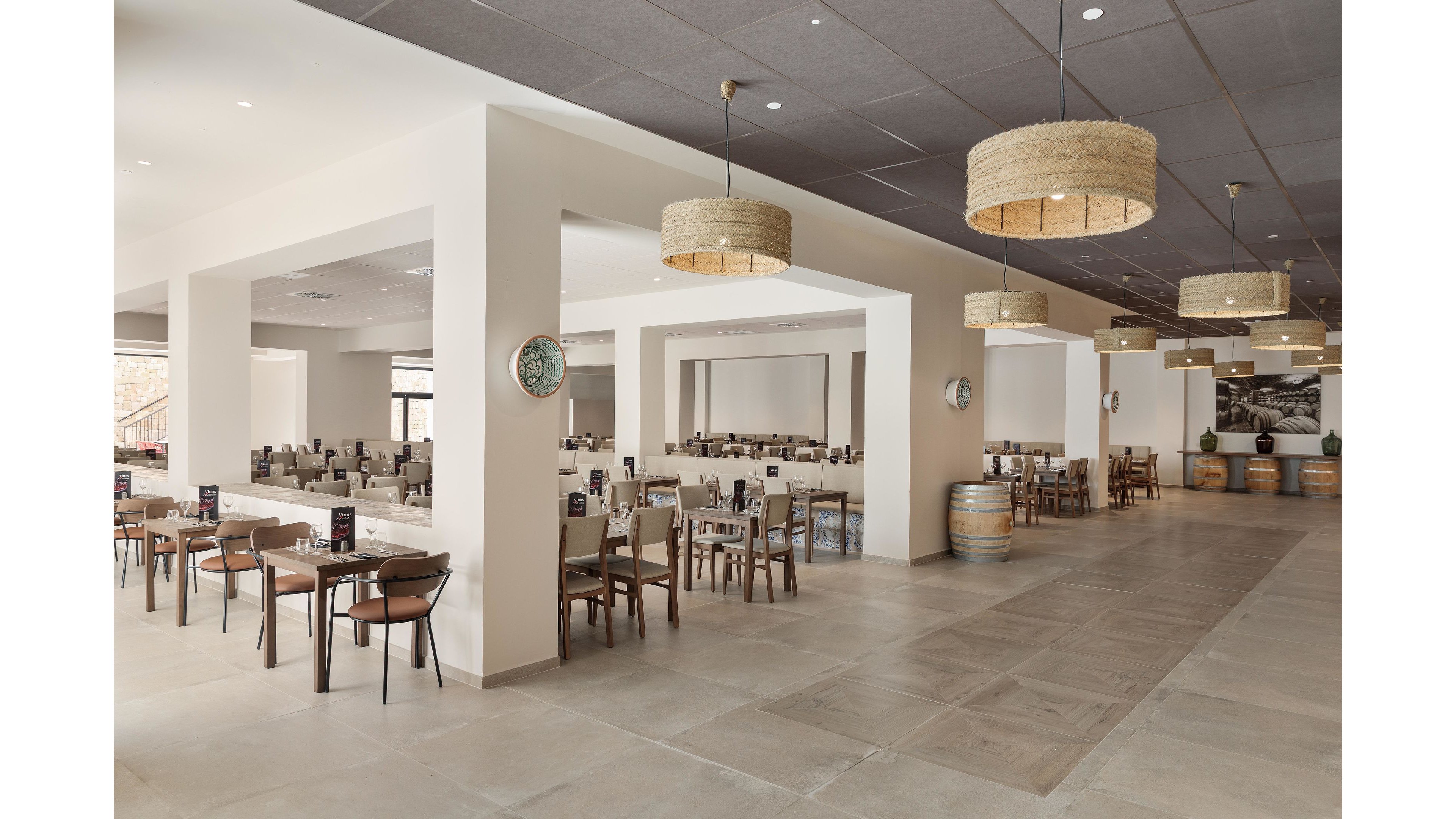 The image shows a large, contemporary restaurant dining area with neatly arranged tables and chairs. The setting features neutral beige and brown tones, woven pendant lights, and decorative barrels, creating a warm and inviting atmosphere. The space is well-lit with natural and artificial lighting, and the overall style is minimalist and modern. No visible text or numeric values are present in the image.
