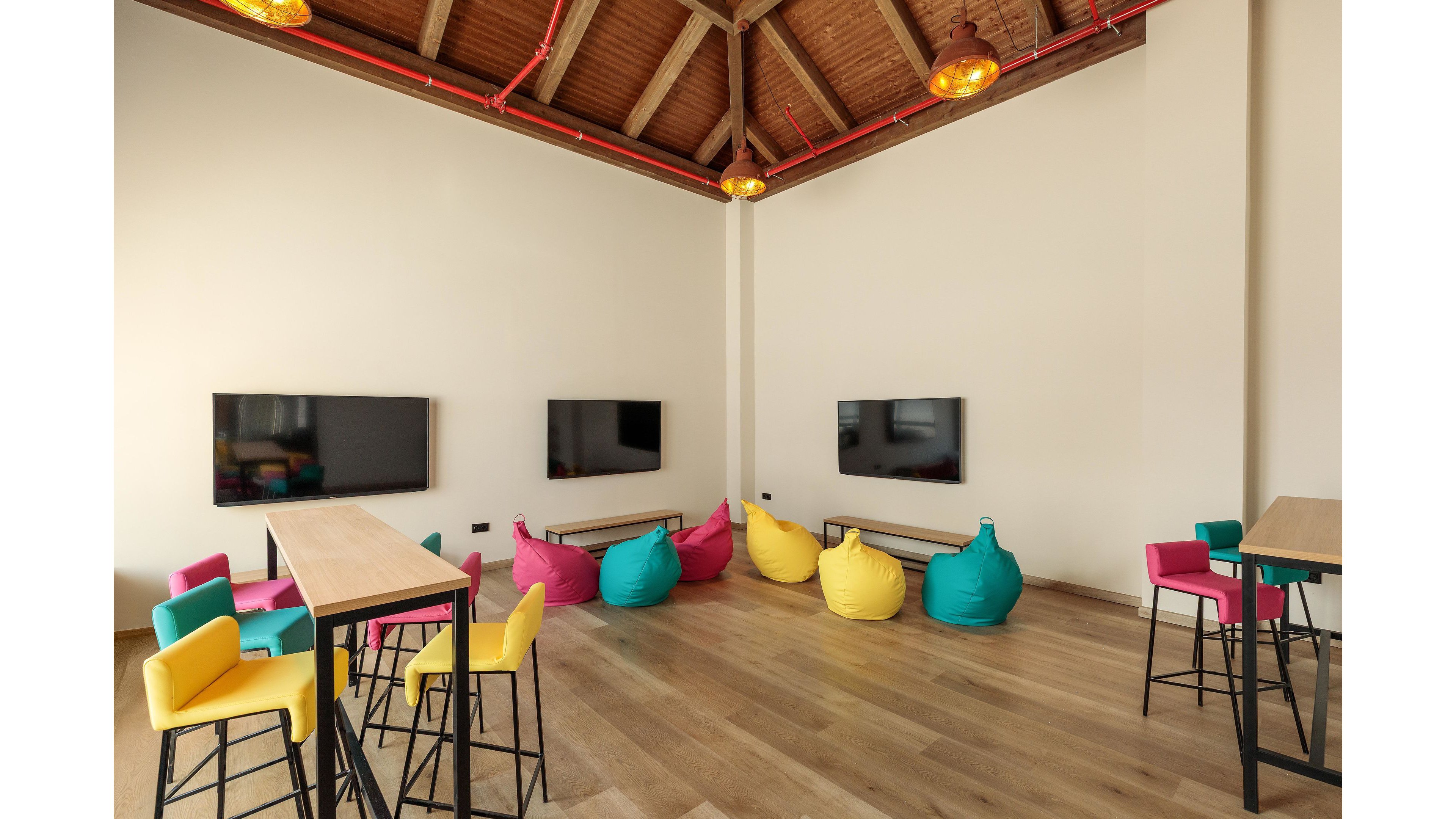 A contemporary lounge area features vibrant bean bag chairs in teal, yellow, and pink, paired with matching bar stools. Three large flat-screen monitors are mounted on the walls, creating a tech-friendly environment. The space is well-lit with warm pendant lights and has a wooden ceiling and floor, giving it a cozy yet modern feel. No visible text or numeric values are present in the image.
