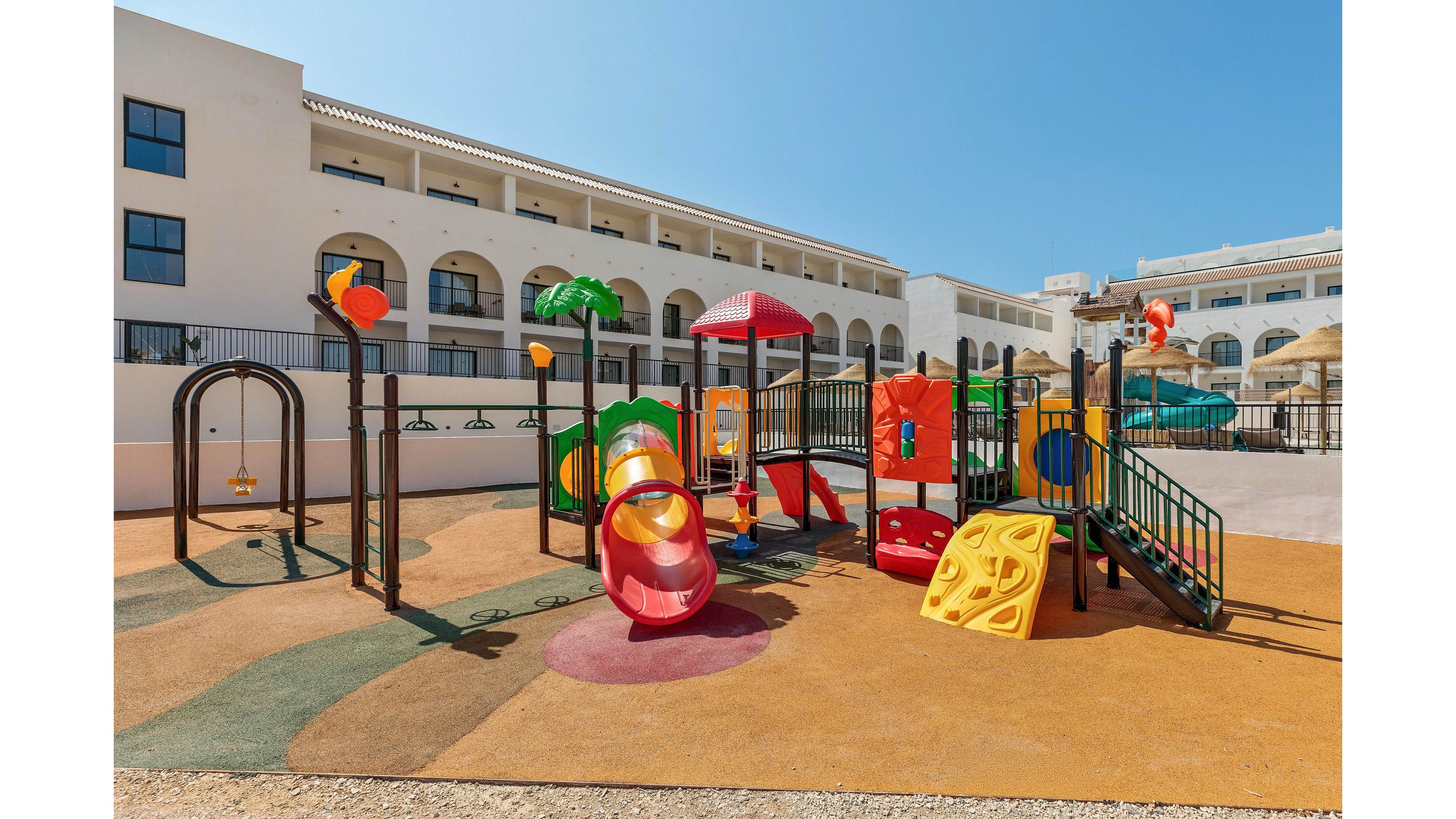 A vibrant children's playground is set outdoors beside a large, white hotel or apartment complex. The play area features slides, tunnels, climbing structures, and swings, all in bright colors like red, yellow, and green. The ground is covered with soft, patterned safety flooring, and the setting is sunny with clear blue skies. No visible text or numbers are present in the image.