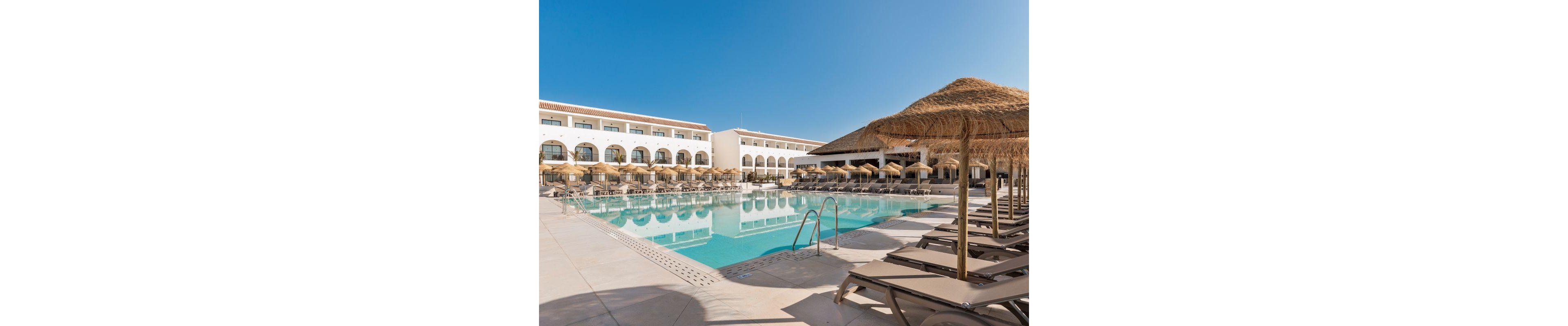 A spacious outdoor swimming pool is surrounded by numerous sun loungers and straw parasols at a modern resort. The setting features a large, white building with arched windows and balconies, reflecting in the clear blue water. The scene is bright and tranquil, with a clear sky and no visible people, evoking a relaxing vacation atmosphere.