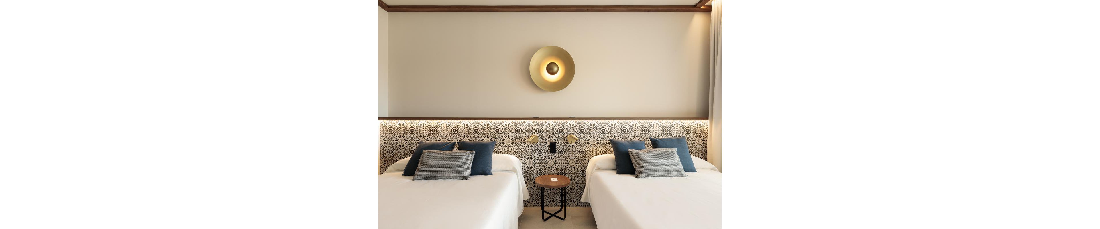 The image shows a contemporary hotel room featuring two neatly made twin beds with white linens and blue accent pillows. The beds are separated by a small round wooden table, and the headboard displays a geometric black-and-white pattern. Above the headboard, a gold circular wall light adds a stylish focal point to the neutral-toned space. The room exudes a calm and inviting atmosphere with its minimalist decor and soft lighting.