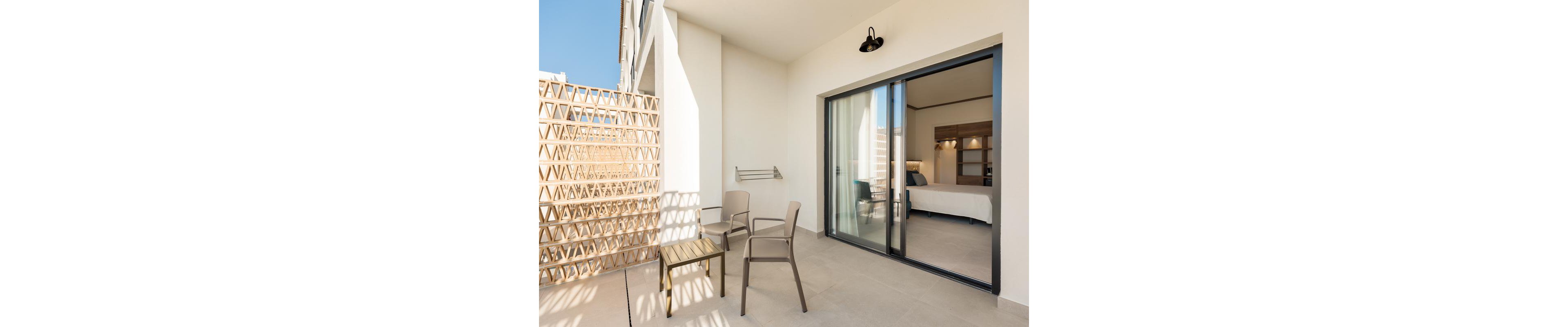 The image shows a contemporary hotel balcony featuring two taupe chairs and a small table. The balcony is enclosed by a geometric privacy screen and opens to a bedroom through large sliding glass doors. The setting is bright and minimal, with neutral tones and clean lines, creating a calm and inviting atmosphere.